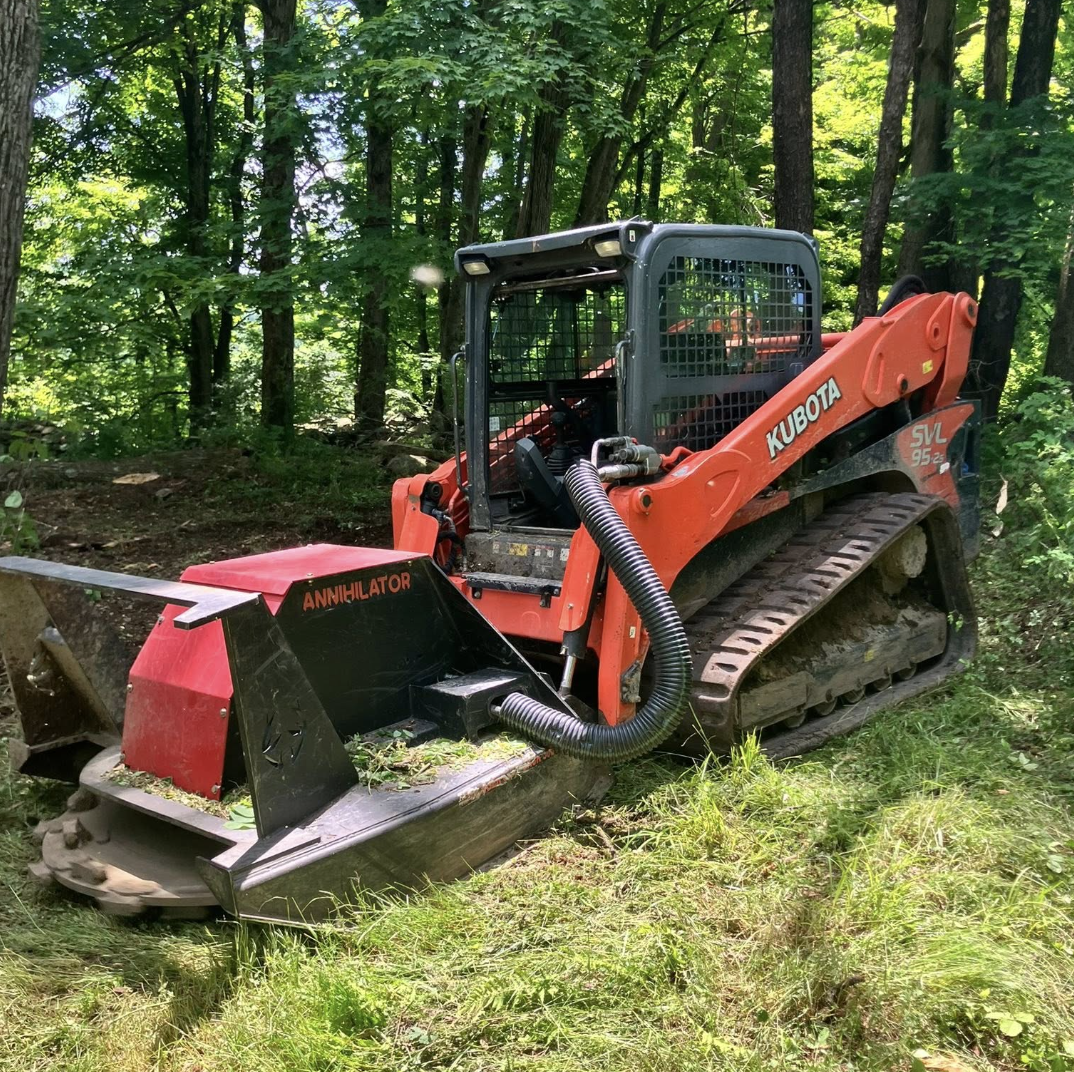 A Kubota tracked forestry mulcher with an orange body and black attachment labeled 'Annihilator' on a grassy clearing surrounded by tall trees.