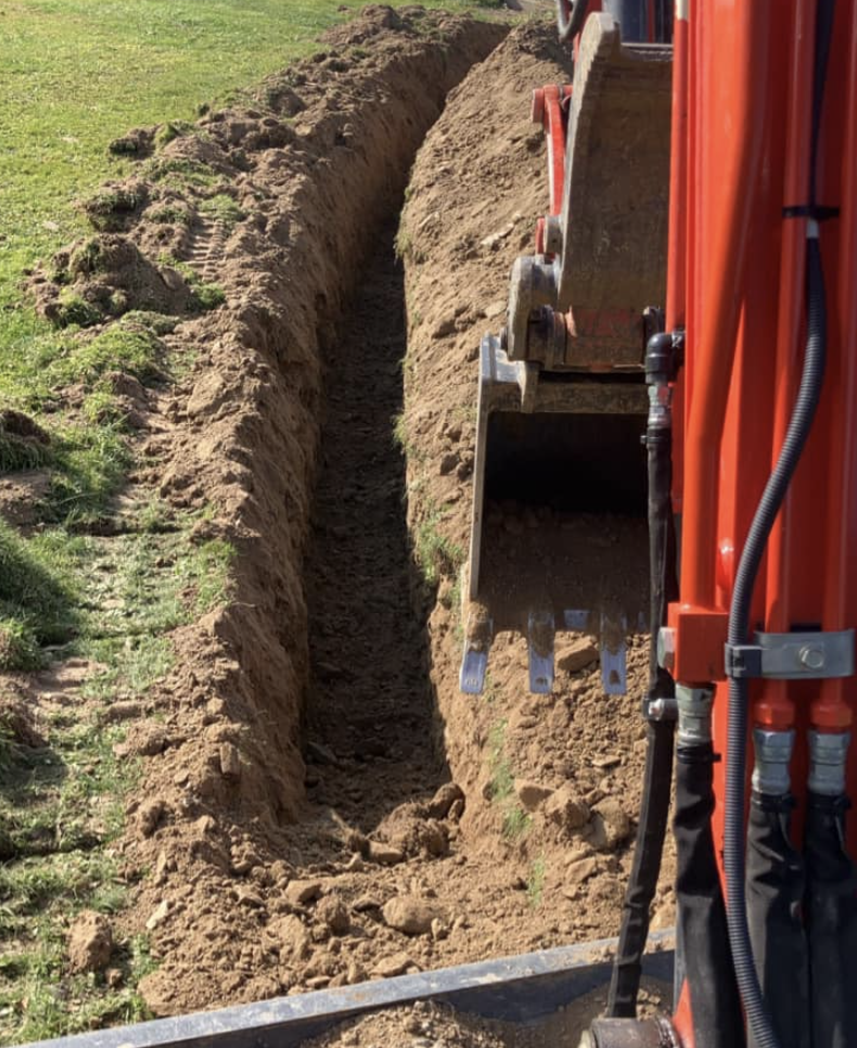 A narrow trench being dug in a grassy area using an excavator with orange and black parts.