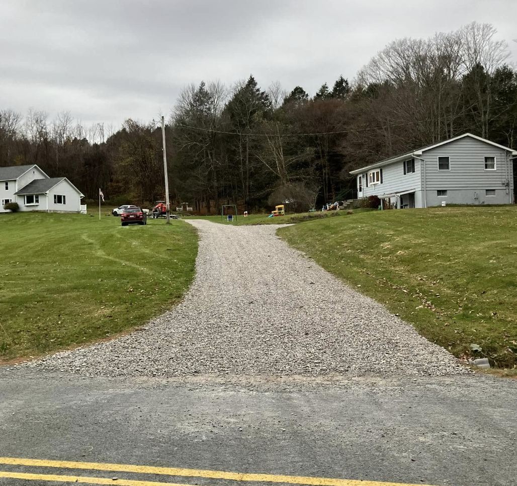 A gravel driveway leading uphill in a suburban neighborhood with green lawns and houses on both sides, and a wooded hillside in the background under an overcast sky.