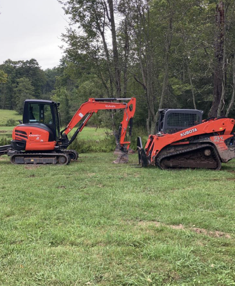 Two orange Kubota compact excavators working together on a grassy field near a wooded area.