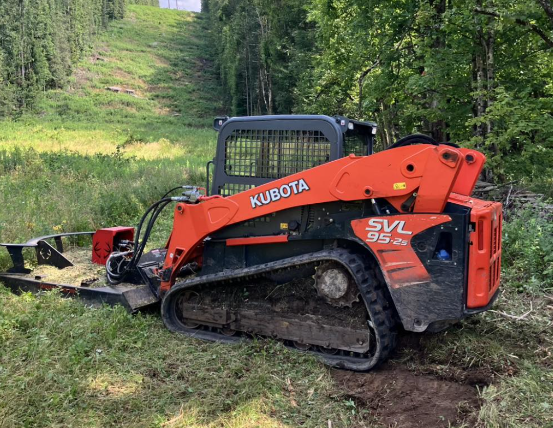 A small Kubota track loader parked on a grassy area near a forested hillside.