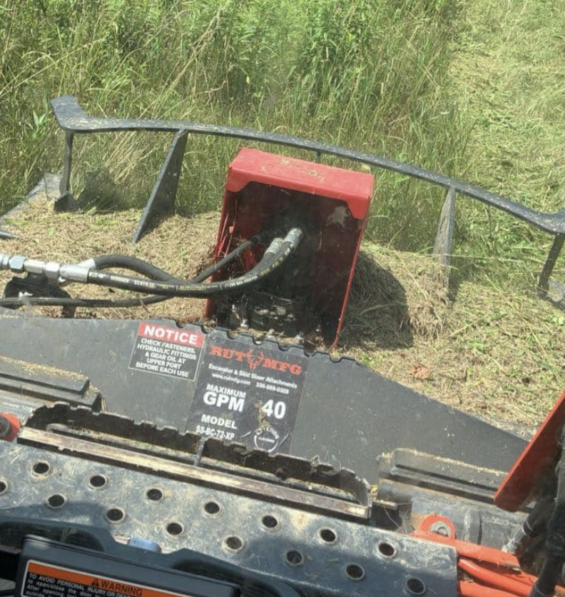A close-up view of a piece of lawn equipment, possibly a mower or trimming attachment, next to tall grass, with hydraulic hoses connected to a red motor or pump unit.