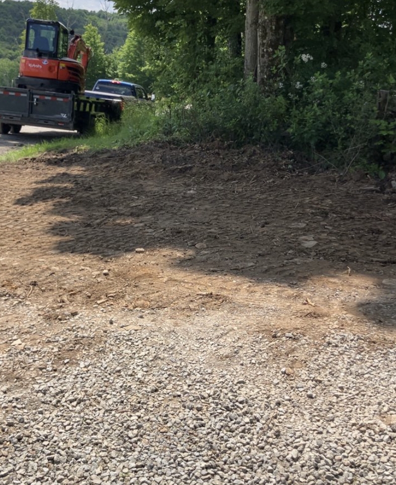 Dirt and gravel path bordered by greenery, with construction equipment and a vehicle on the side of a road in the background.