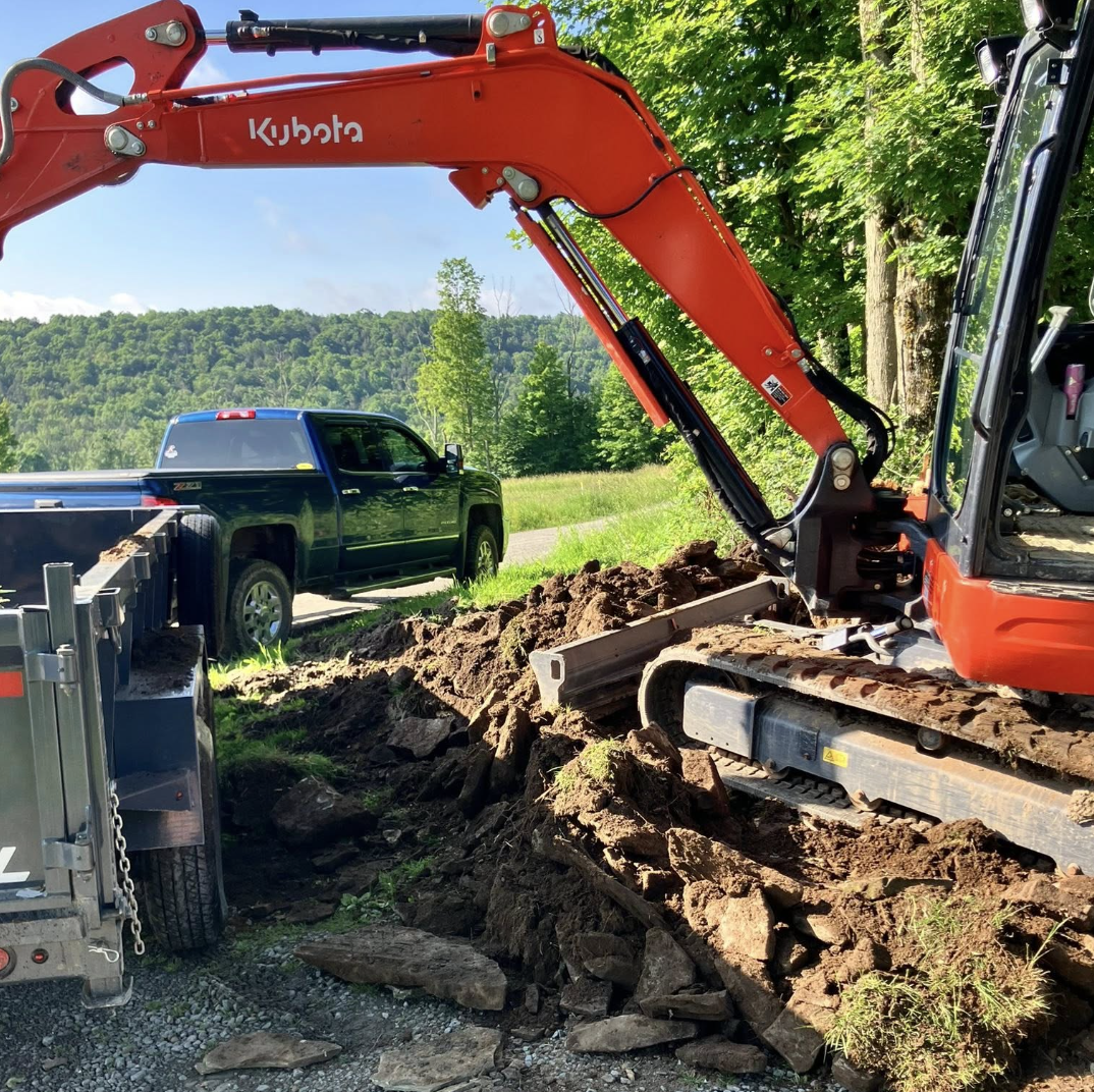 A construction excavation site with an orange Kubota excavator digging up dirt and rocks, and a black pickup truck parked nearby amidst green trees and hills.
