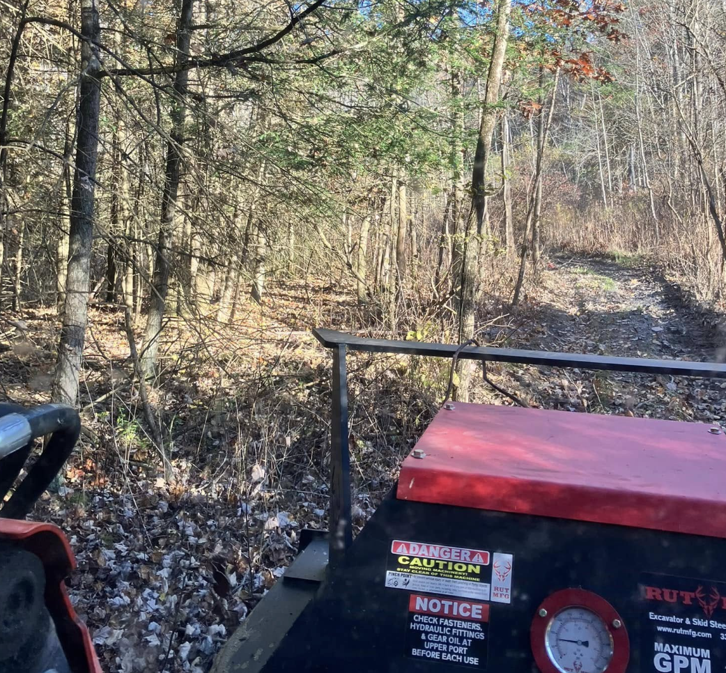 View from a vehicle or machinery on a dirt trail through a wooded forest with trees and fallen leaves.