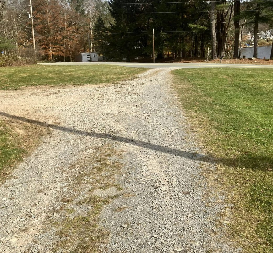 A gravel driveway splits and leads to a paved road in a rural area with trees and a body of water in the background.