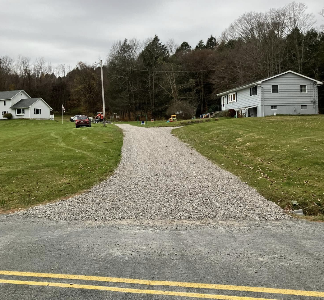 Gravel driveway leading away from a paved road, flanked by green lawns and houses, with trees in the background under an overcast sky.
