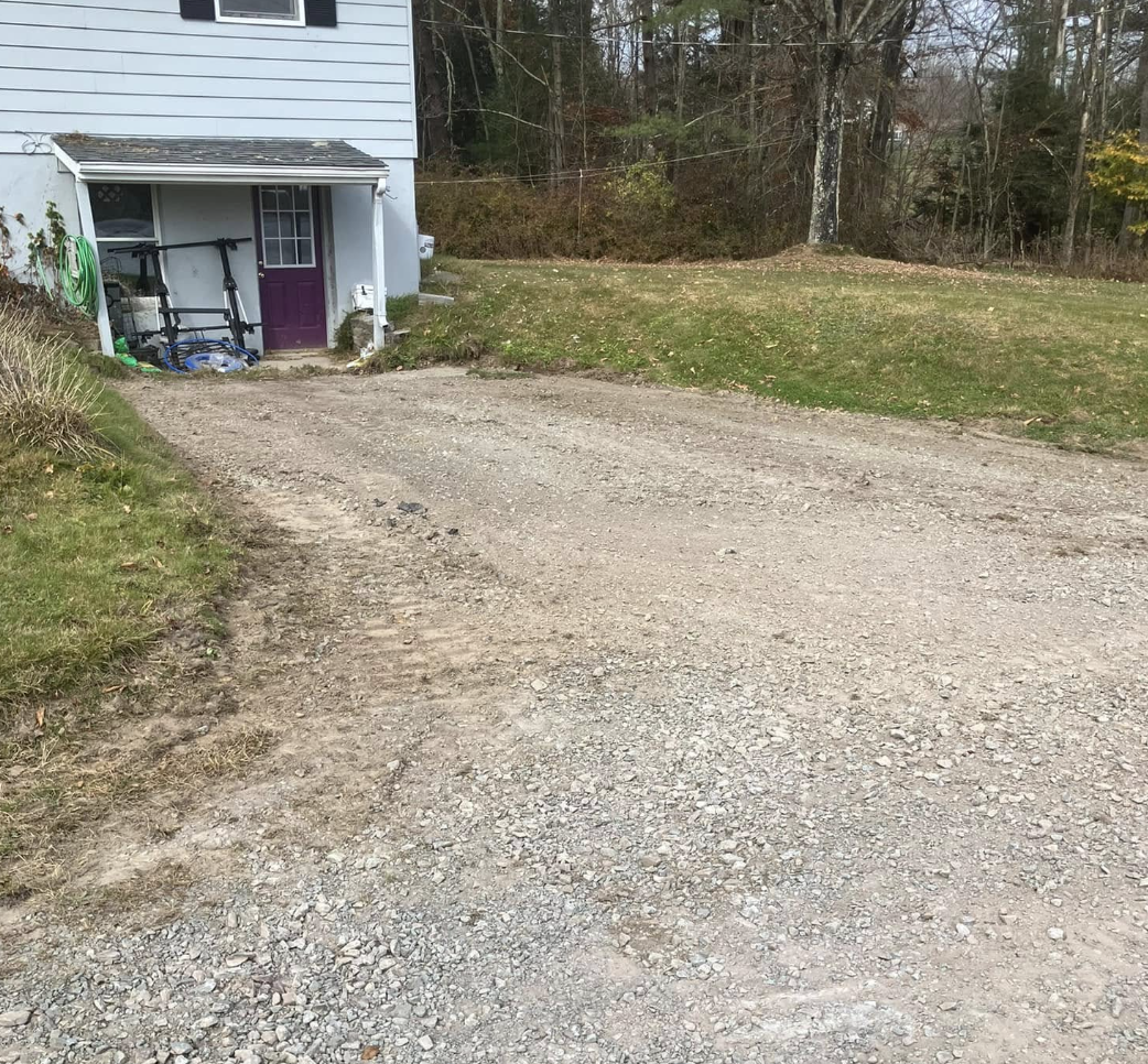 A gravel driveway in front of a house with a purple door, a green hose coiled on the side, and a bicycle leaning against the house. There are trees and a grassy area in the background.