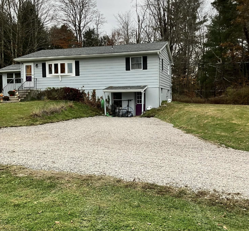 A two-story house with light gray siding, black shutters, and a small covered area in the driveway with outdoor furniture and gardening tools, surrounded by a grassy yard and wooded area in the background.