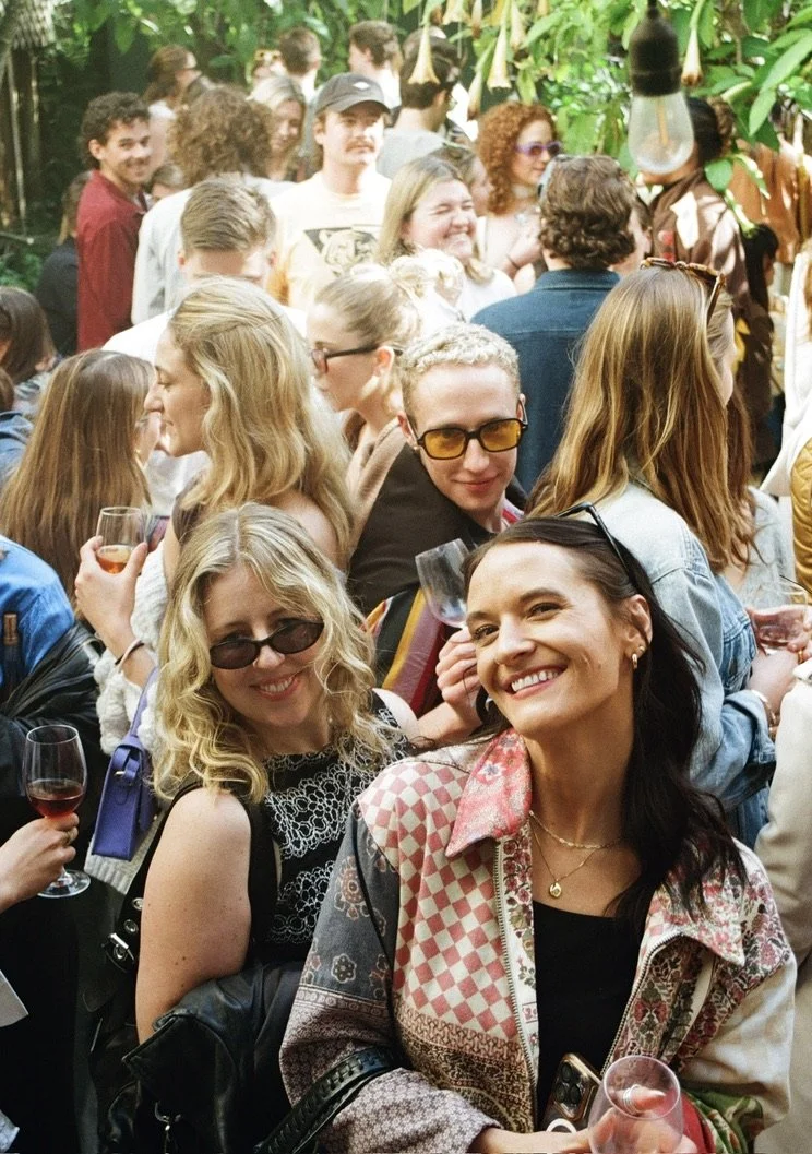 A group of people at a social gathering outdoors, surrounded by green foliage, some holding wine glasses, smiling, and engaging with each other.