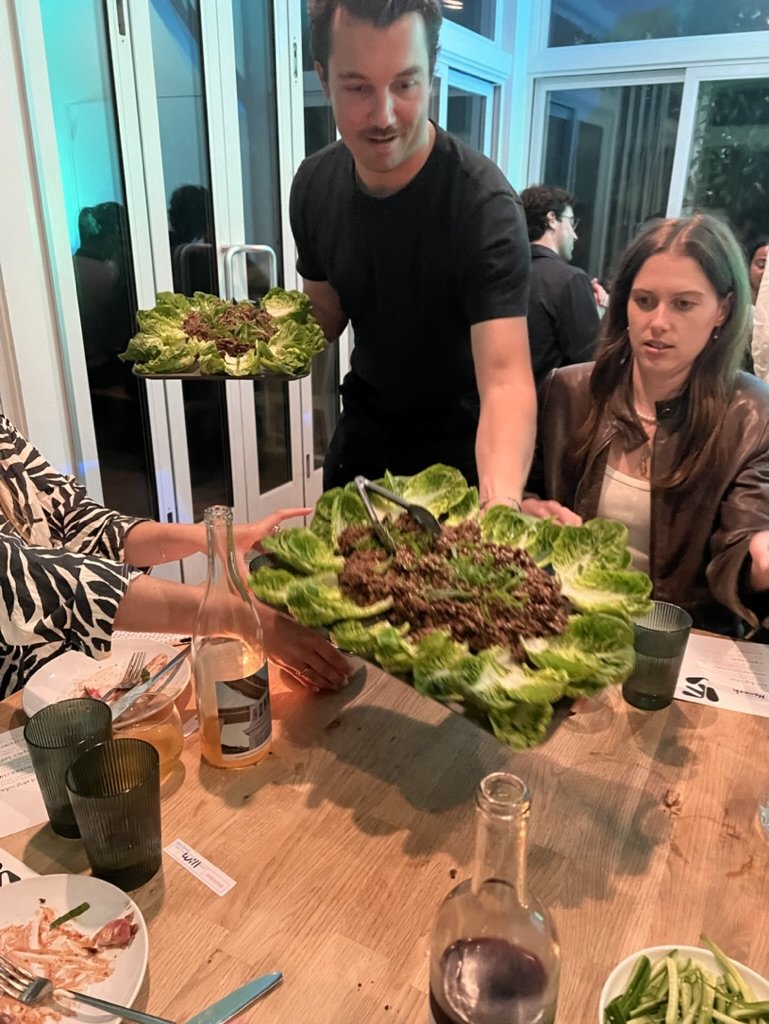Man serving a large platter of salad topped with ground meat and surrounded by romaine lettuce leaves at a dinner table with other guests, Wi-Fi sign and glass bottles on the table.
