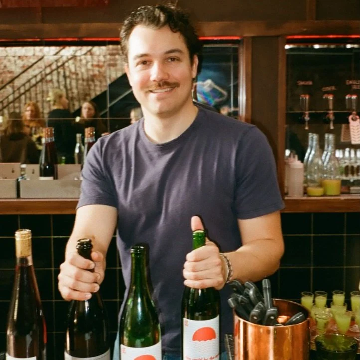 Man with a mustache smiling at camera while holding wine bottles behind a bar with various bottles and glasses in the background.