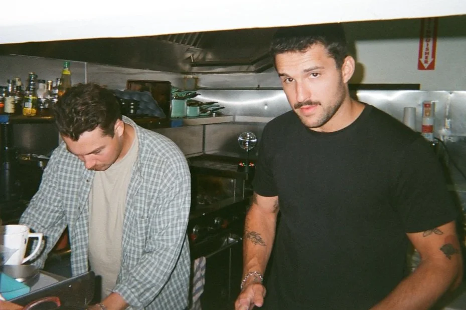 Two men in a kitchen setting, one wearing a checked shirt looking down, and the other wearing a black t-shirt with tattoos on his arms looking at the camera.