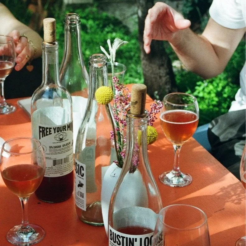 Glass bottles with cork stoppers, some labeled with marijuana references, and glasses of rosé wine on an outdoor table with flowers in a vase, in a green natural setting.