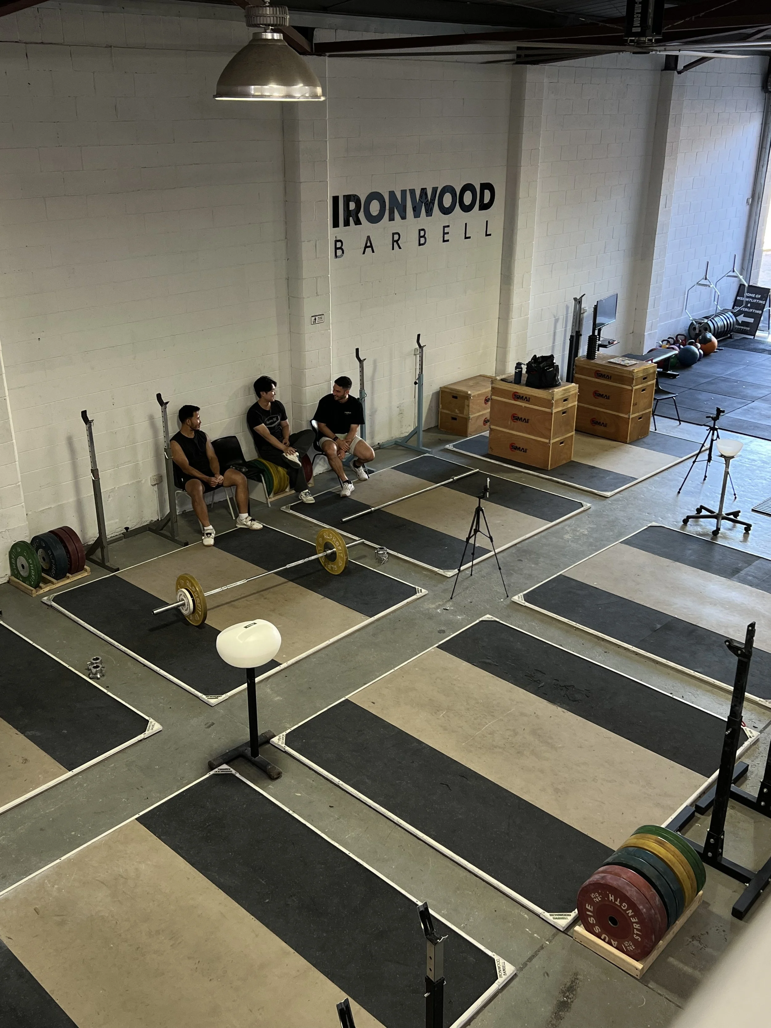 Inside Ironwood Barbell gym with weightlifting platforms, bumper plates, and squat racks, with three people sitting on chairs against the wall.