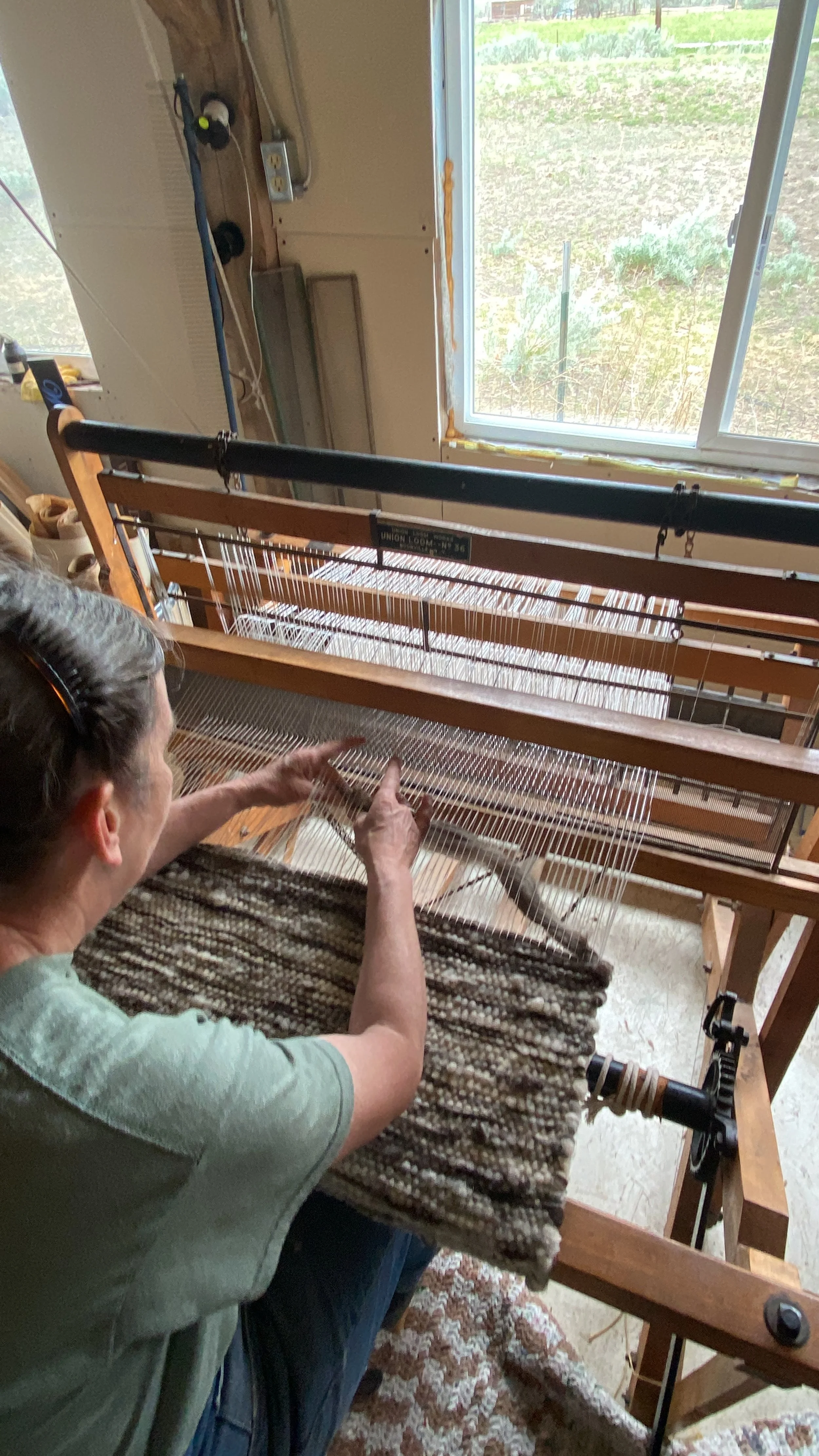 A woman operating a weaving loom indoors, with a window showing grass and plants outside.