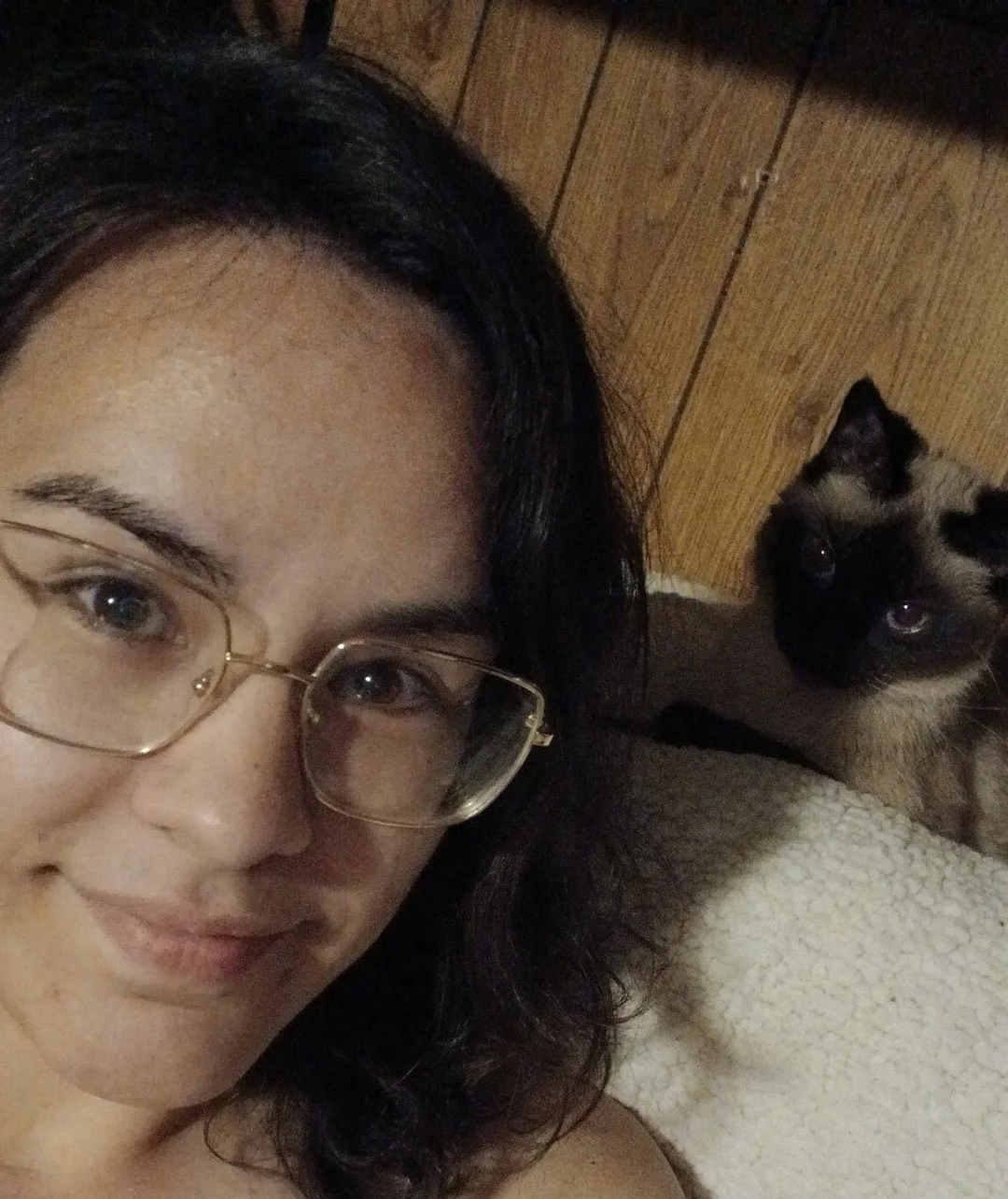 A woman with dark hair and glasses lying on a couch with a Siamese cat nearby on the same couch.
