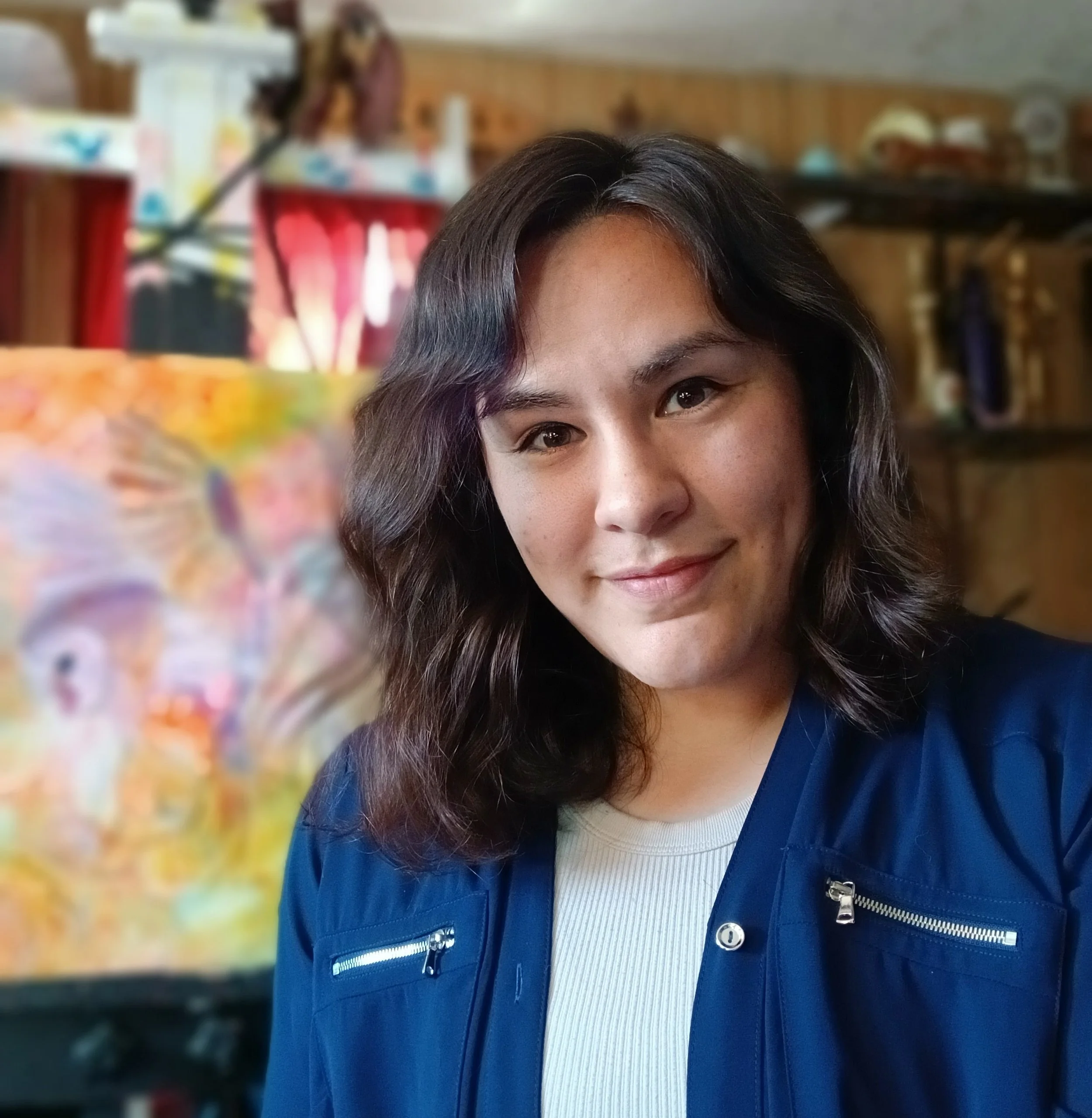 A young woman with shoulder-length wavy dark hair smiling at the camera, wearing a blue jacket over a white shirt, with a blurred background of a wooden-paneled room and colorful artwork.