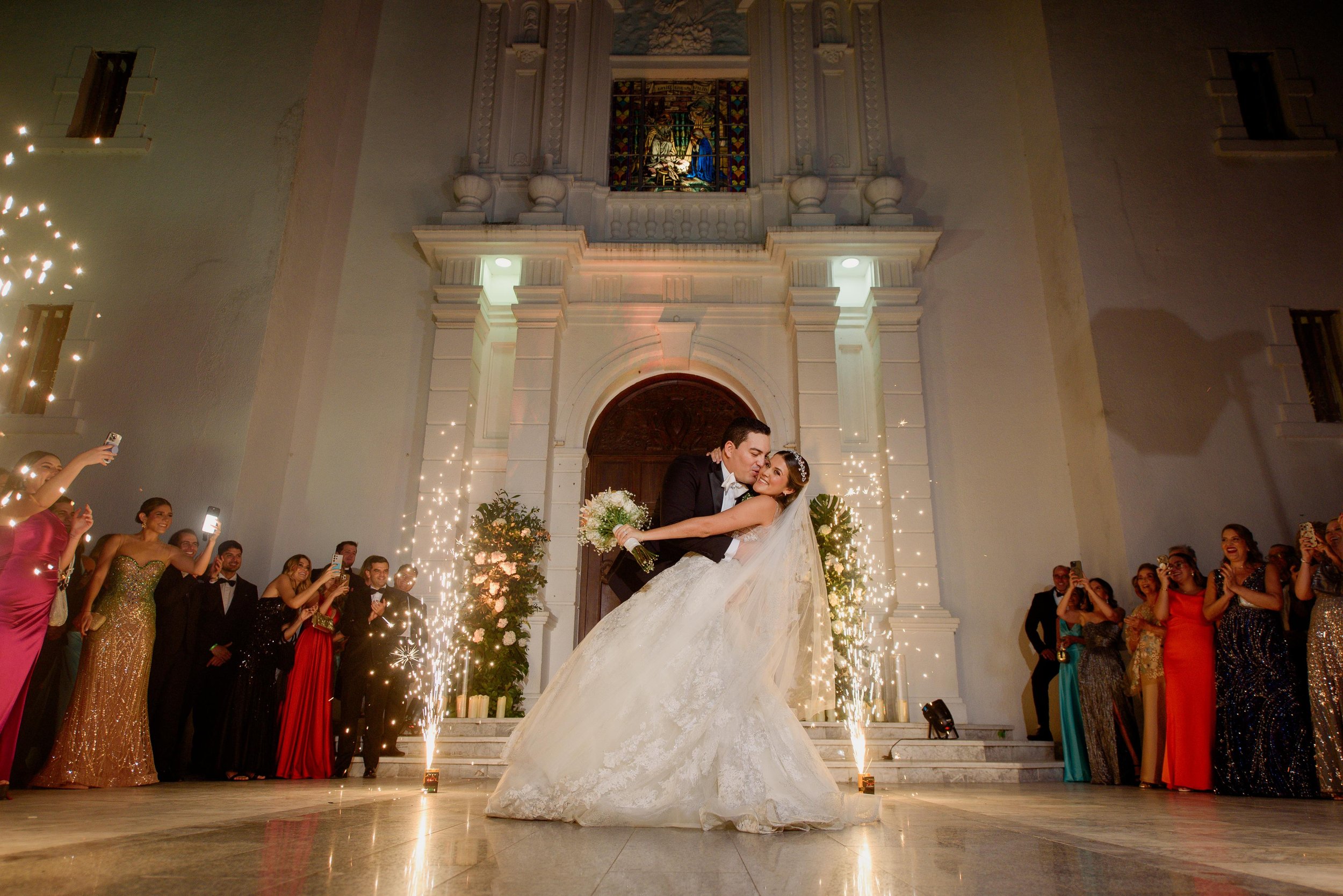 Novios bailando en su boda frente a iglesia, rodeados de invitados y efecto de sparkular.