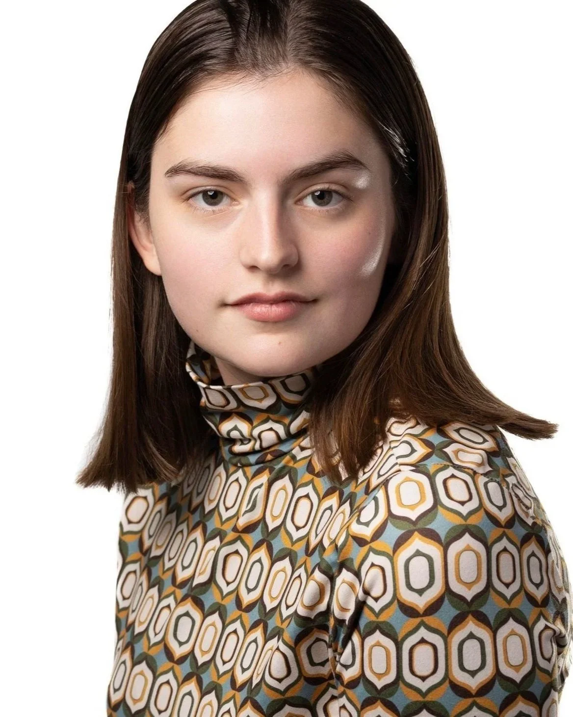 Young woman with shoulder-length brown hair wearing a patterned high-neck shirt, looking directly at the camera with a neutral expression against a white background.