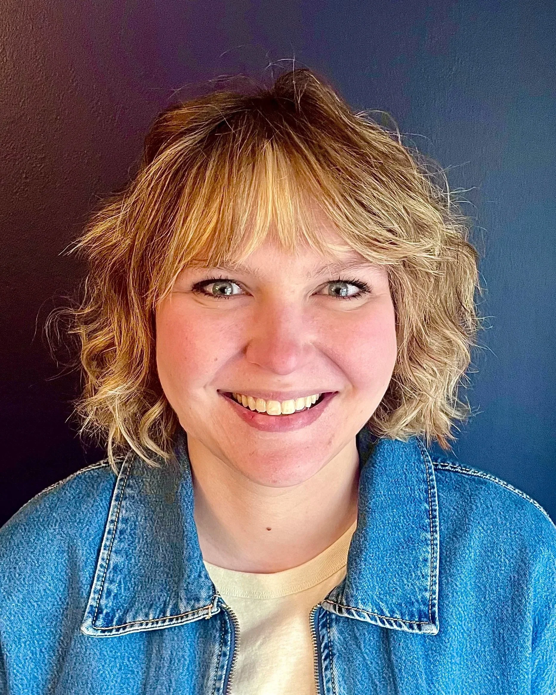 Close-up portrait of woman with curly blonde hair, smiling, wearing a denim jacket over a yellow shirt, with dark background.