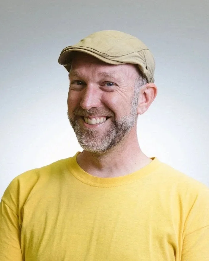 A smiling middle-aged man with a beard, wearing a yellow T-shirt and a beige flat cap, standing against a plain light-colored background.