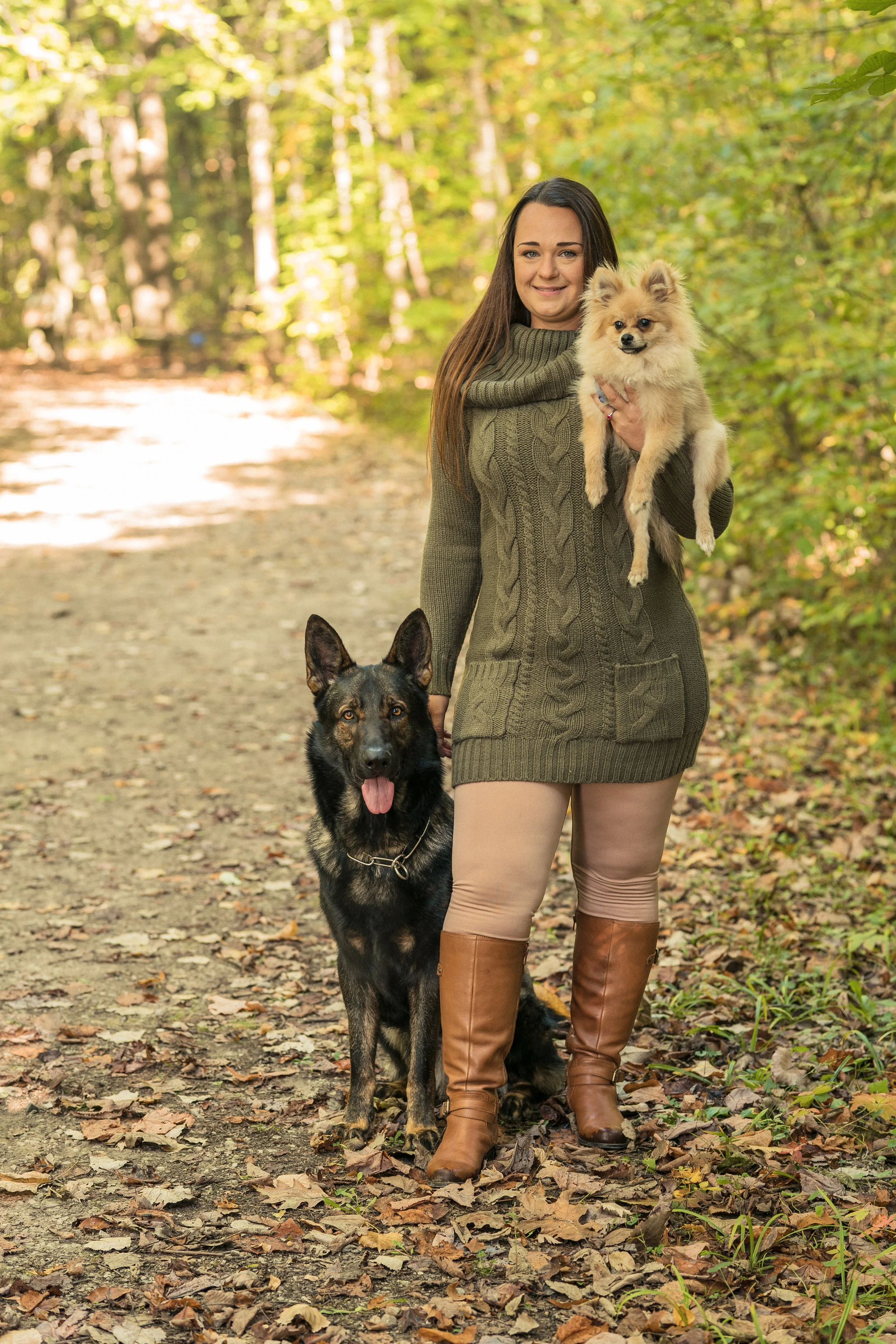 Amanda Cox, the dog trainer at The Possible K9, holding a Pomeranian and standing with a German Shepherd on a forest trail in autumn.