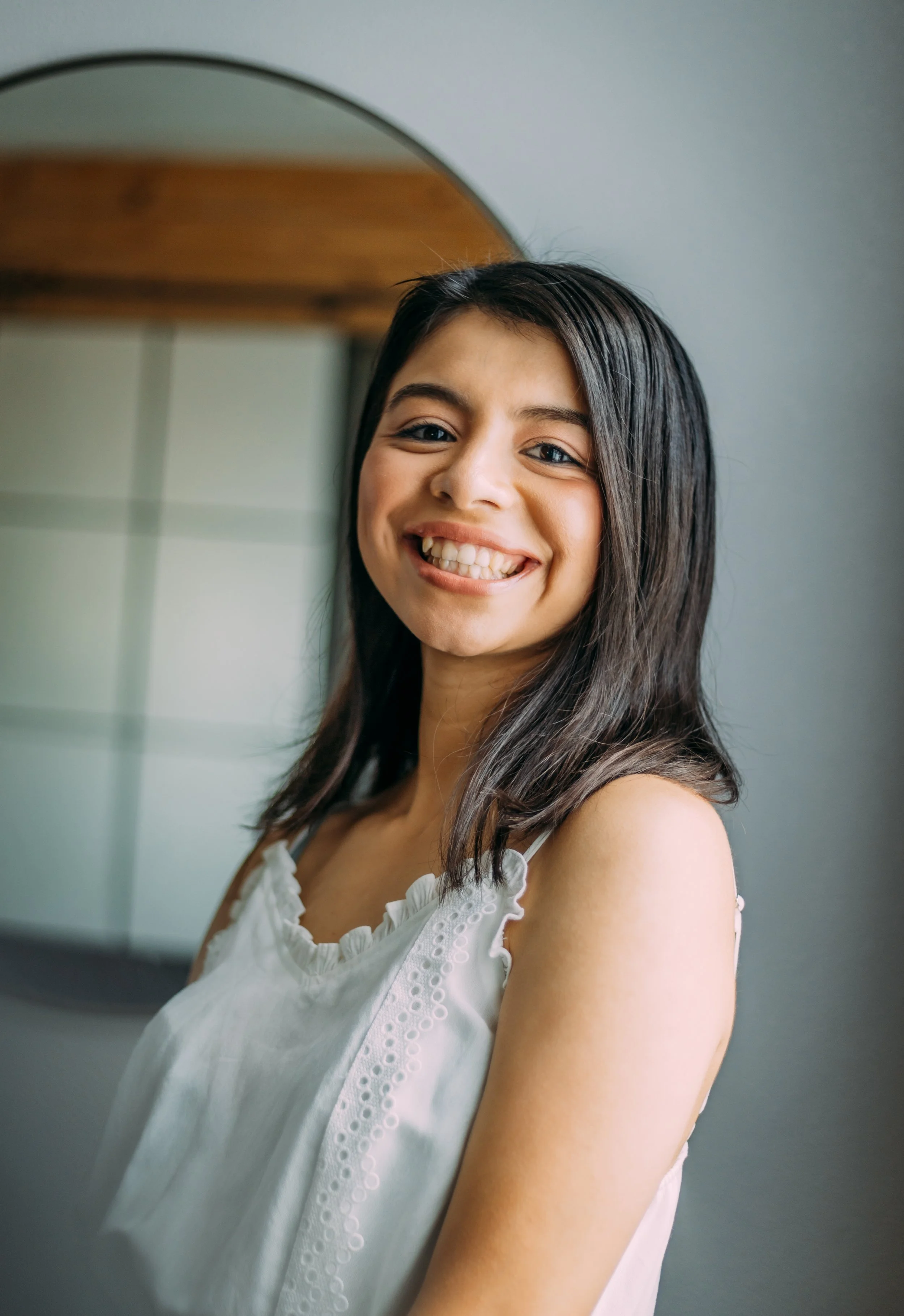 Young woman smiling, looking at mirror in a room with a round mirror and kitchen cabinets in the background.