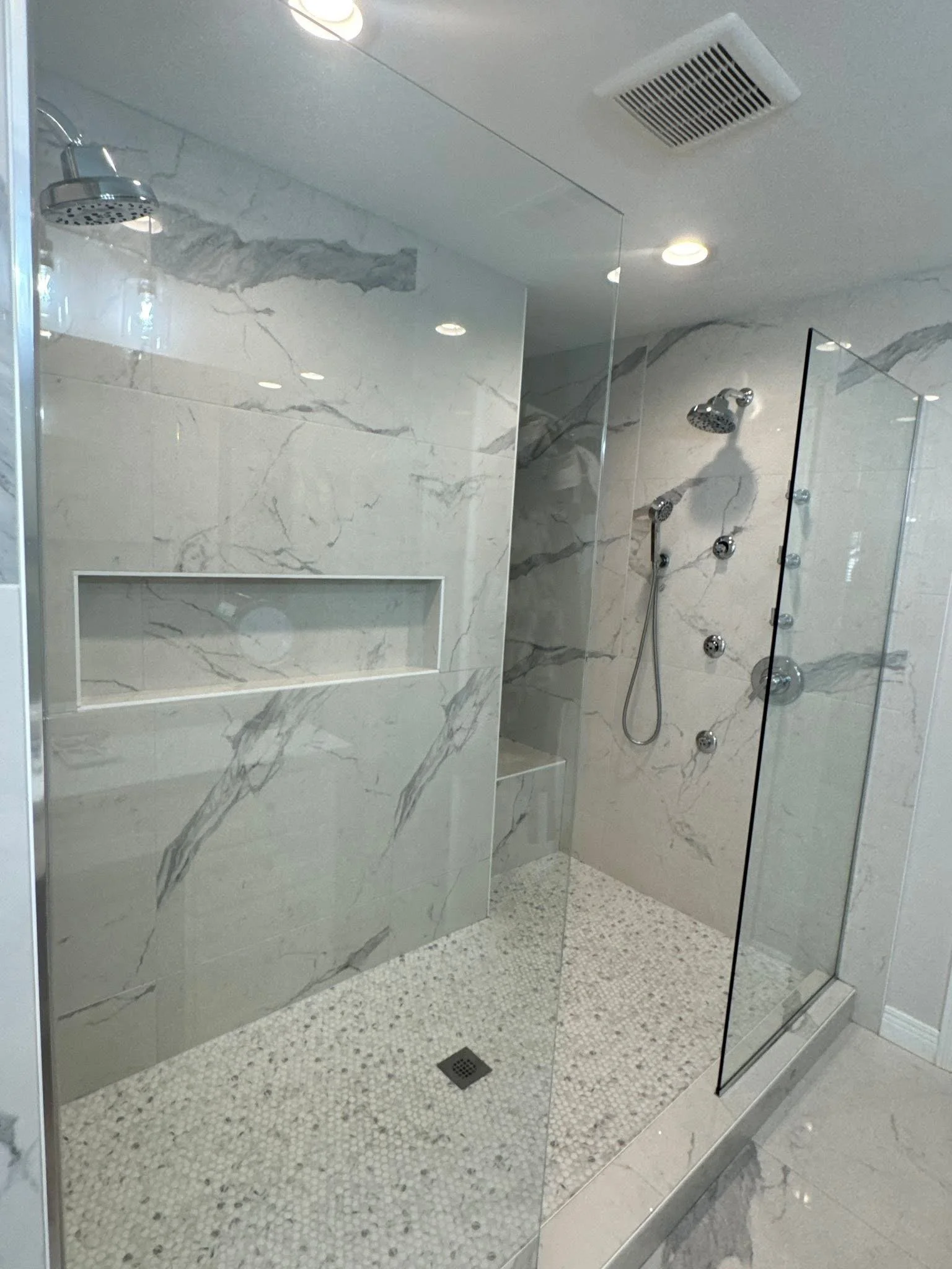 Modern walk-in shower with marble walls, built-in niche, pebble floor, multiple showerheads, and a glass partition.