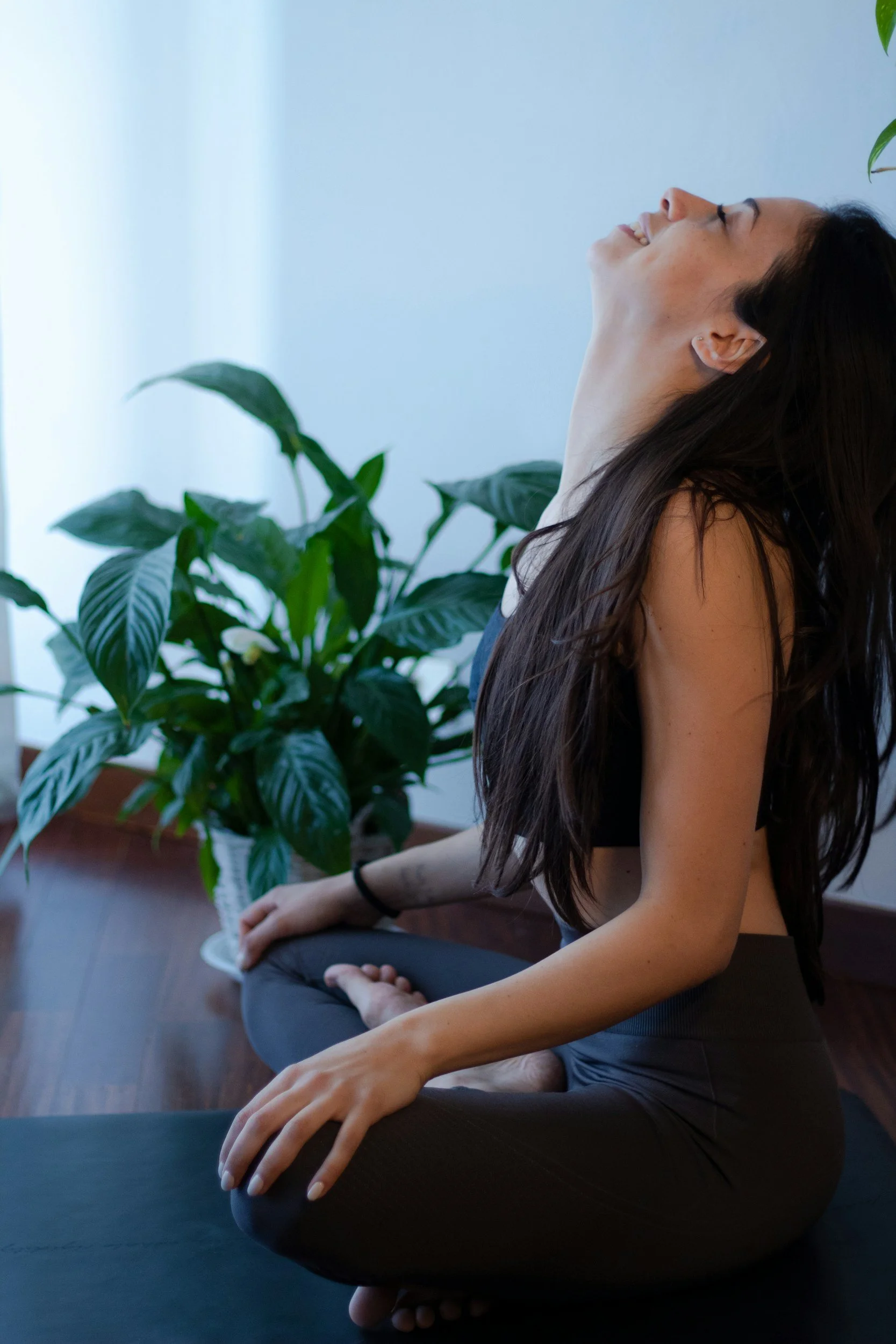 A woman practicing yoga in a cross-legged seated pose, smiling with her head tilted back, near a potted plant in a bright room.