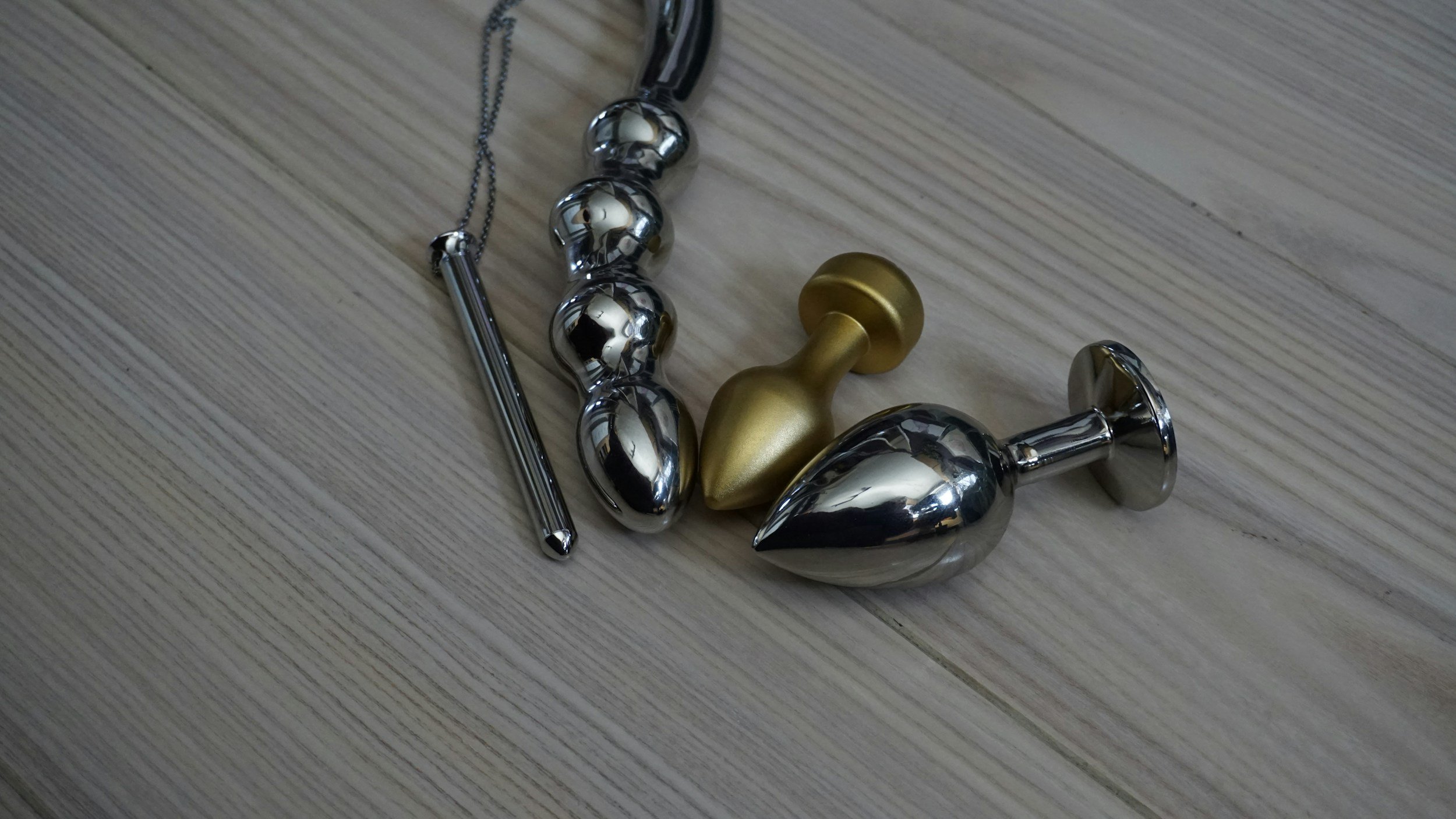 Silver and gold metal anal plugs and a sex toy on a wooden floor.