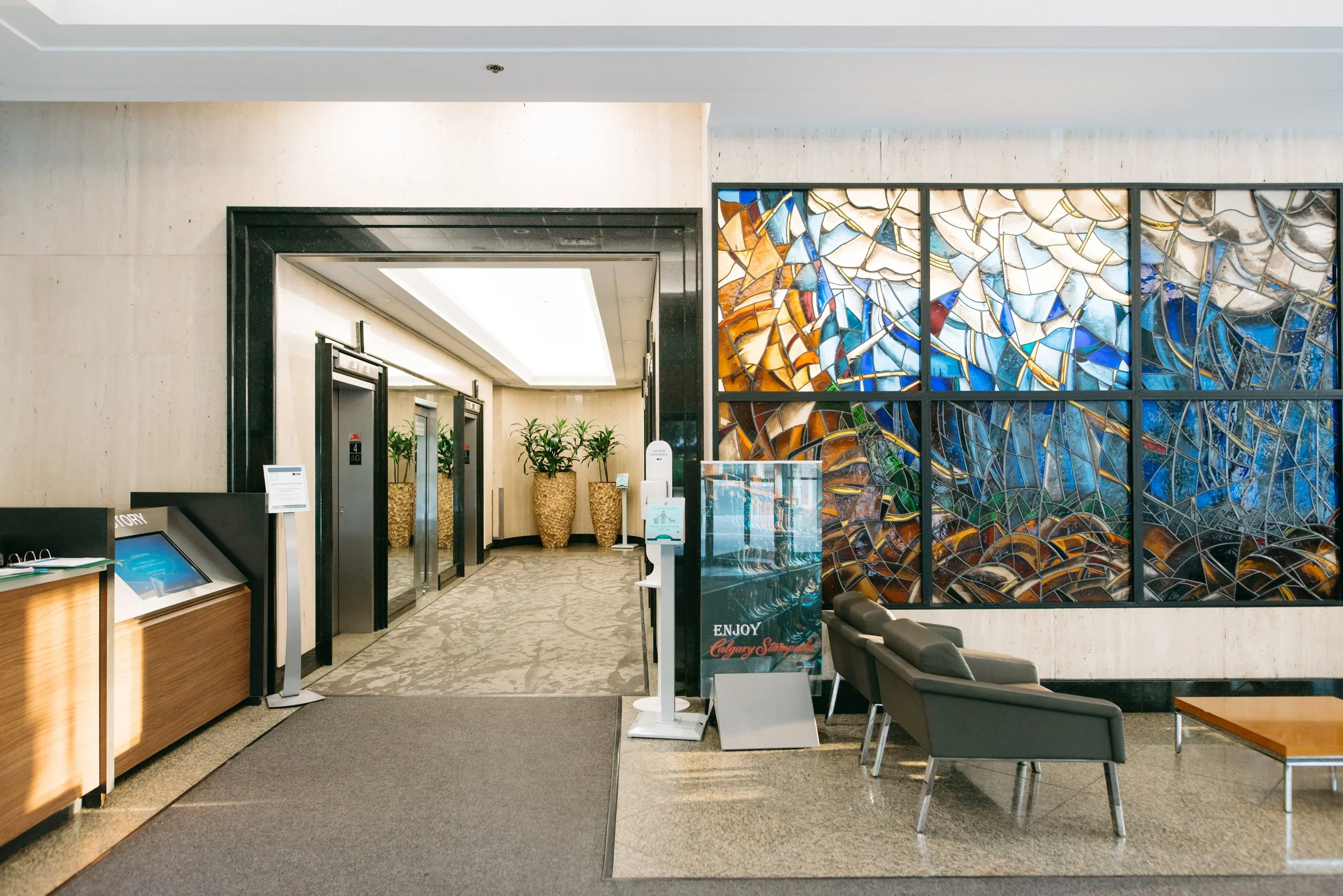 Hotel lobby with elevator doors, seating area with chairs, decorative stained glass wall, potted plants, and signage.