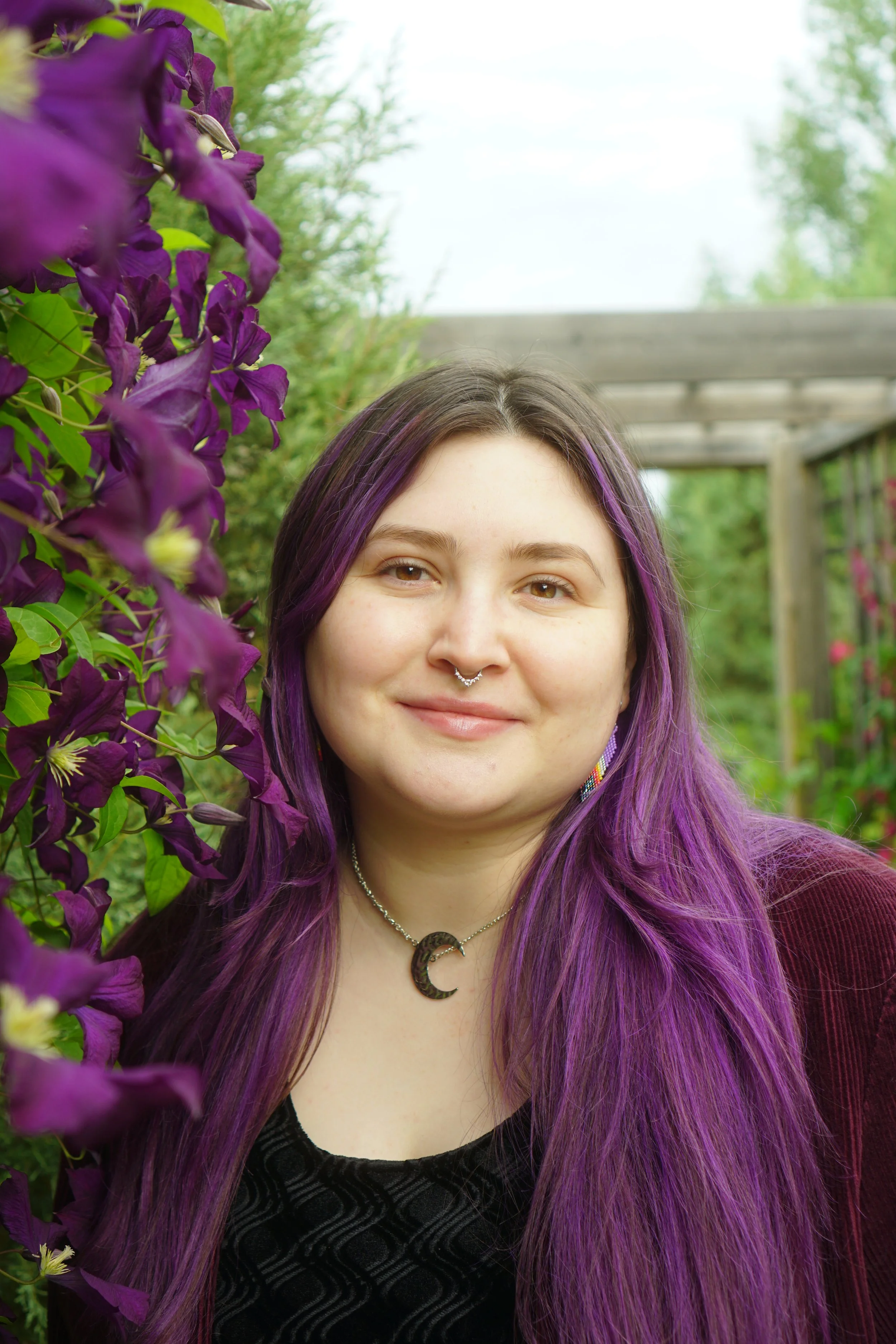 A young woman with long purple hair smiling outdoors, surrounded by purple flowers and greenery.