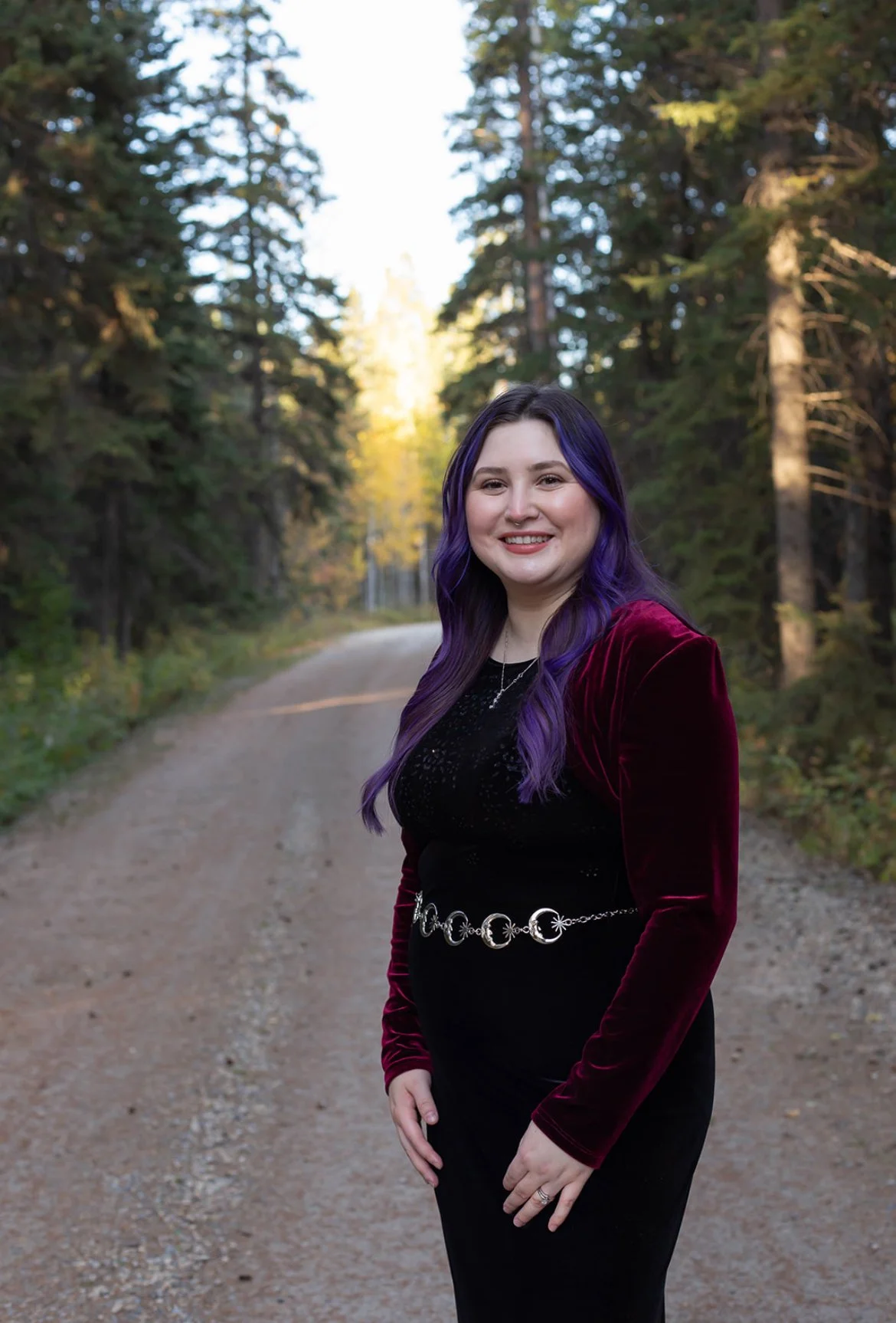A woman with purple hair smiling outdoors on a dirt path surrounded by trees in a forest.