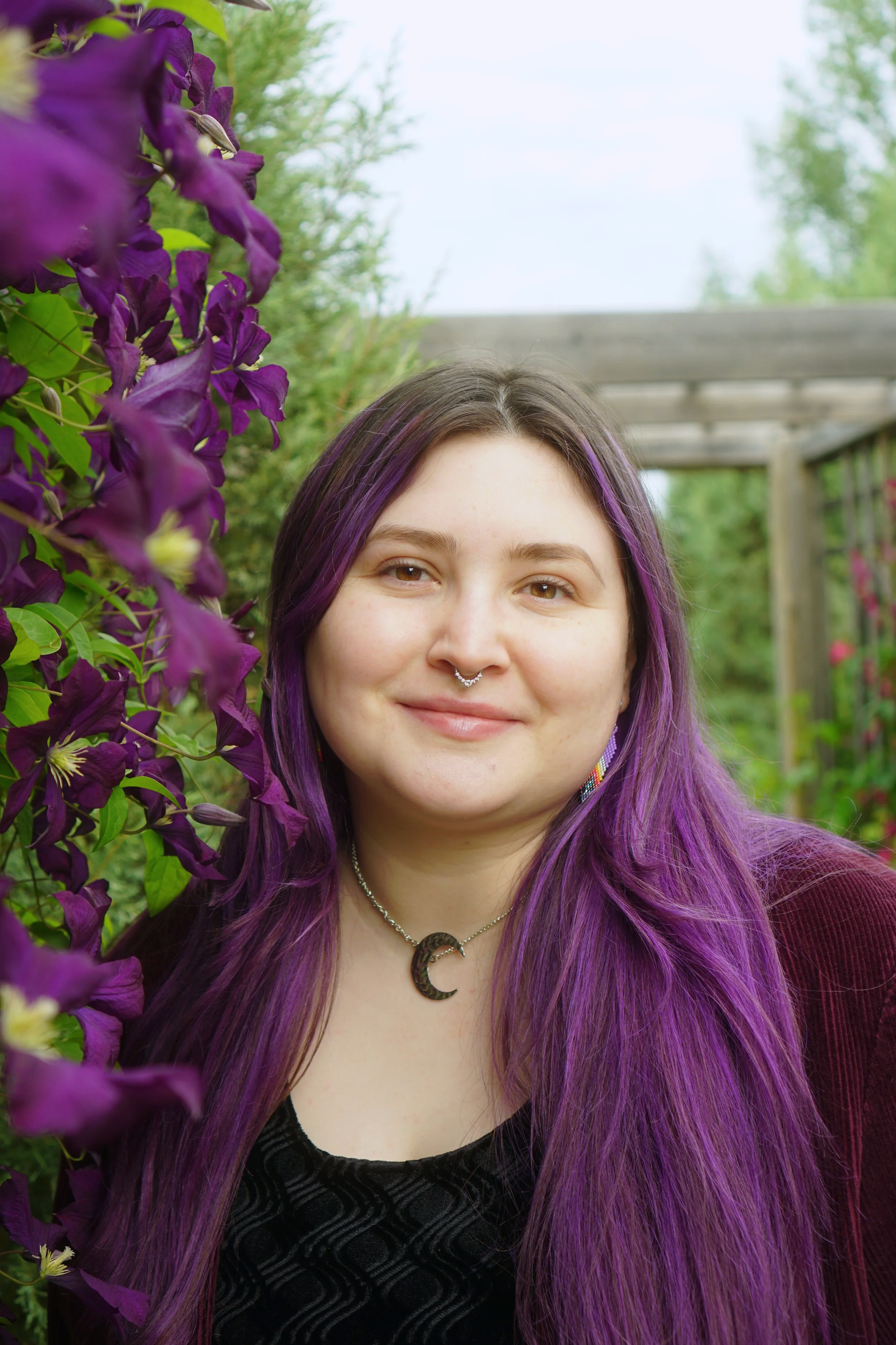 A young woman with purple hair and a septum piercing smiling outdoors next to a purple flowering plant.