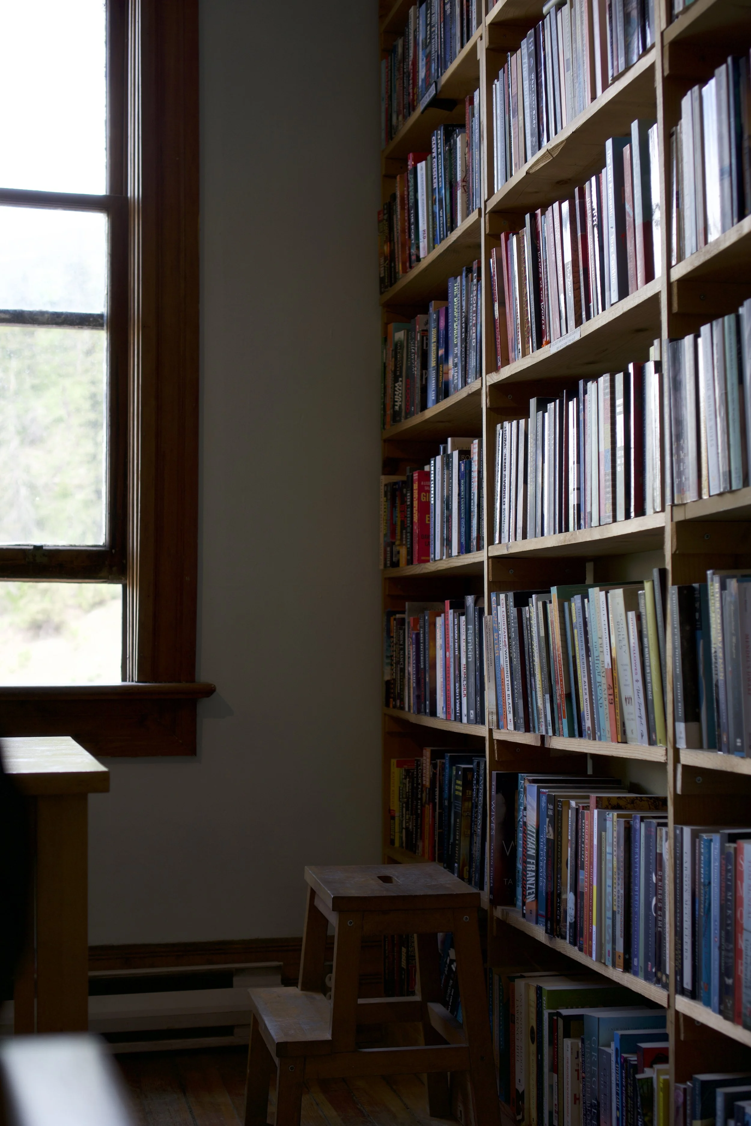A bookshelf filled with books, a window with natural light, and a small wooden stool in a cozy room.