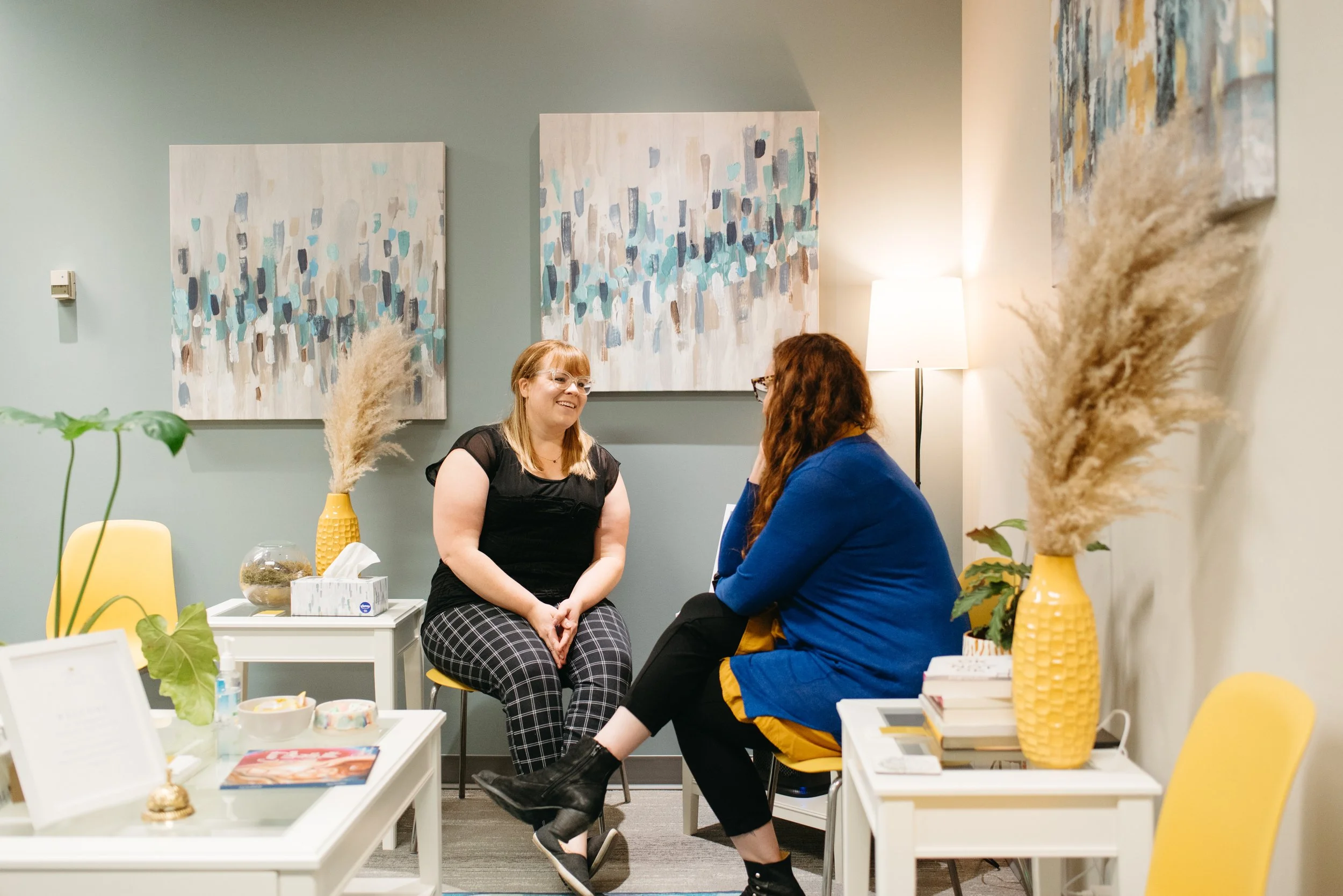 Two people sitting and holding hands, one wearing a denim jacket and the other in black clothing, in a dark setting.