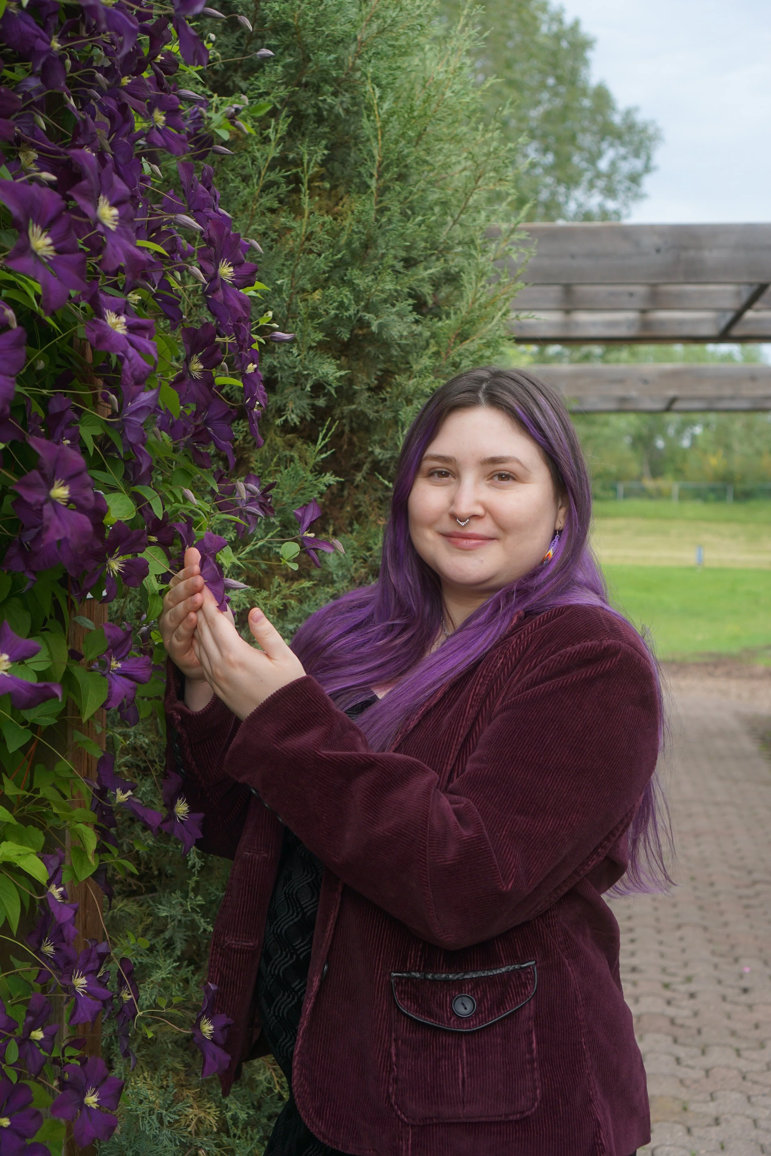 A woman with purple hair and a septum piercing standing outdoors next to a purple flowering plant and green trees, smiling.