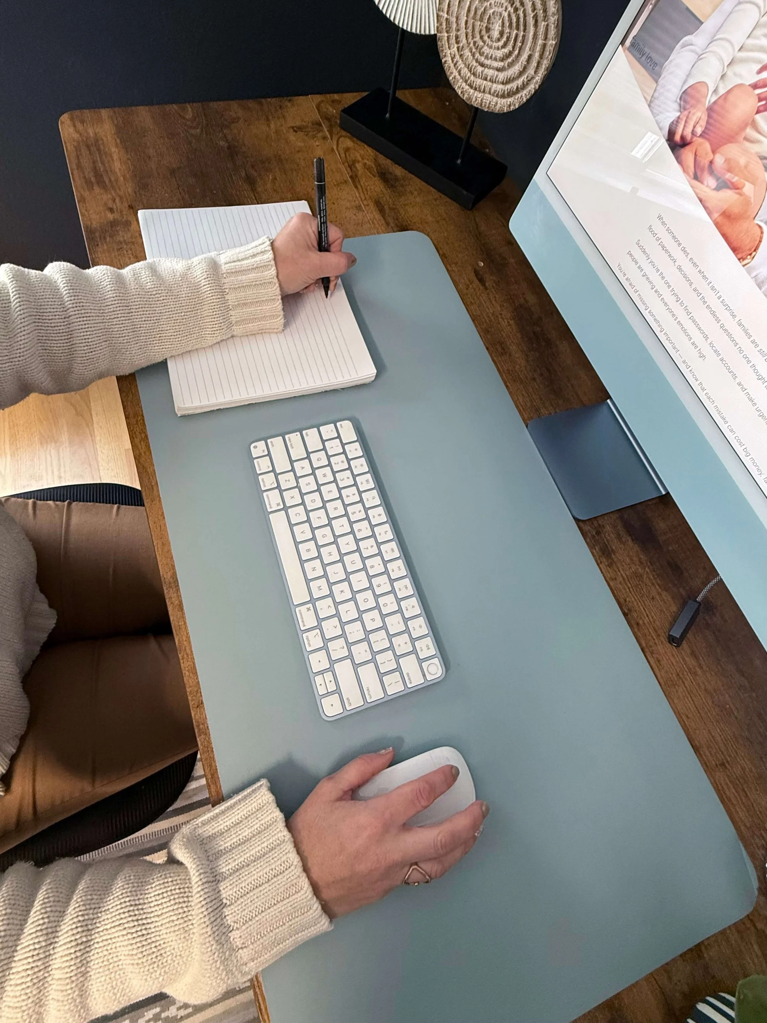 Person using a computer mouse and writing in a notebook on a wooden desk with a keyboard and a large monitor.
