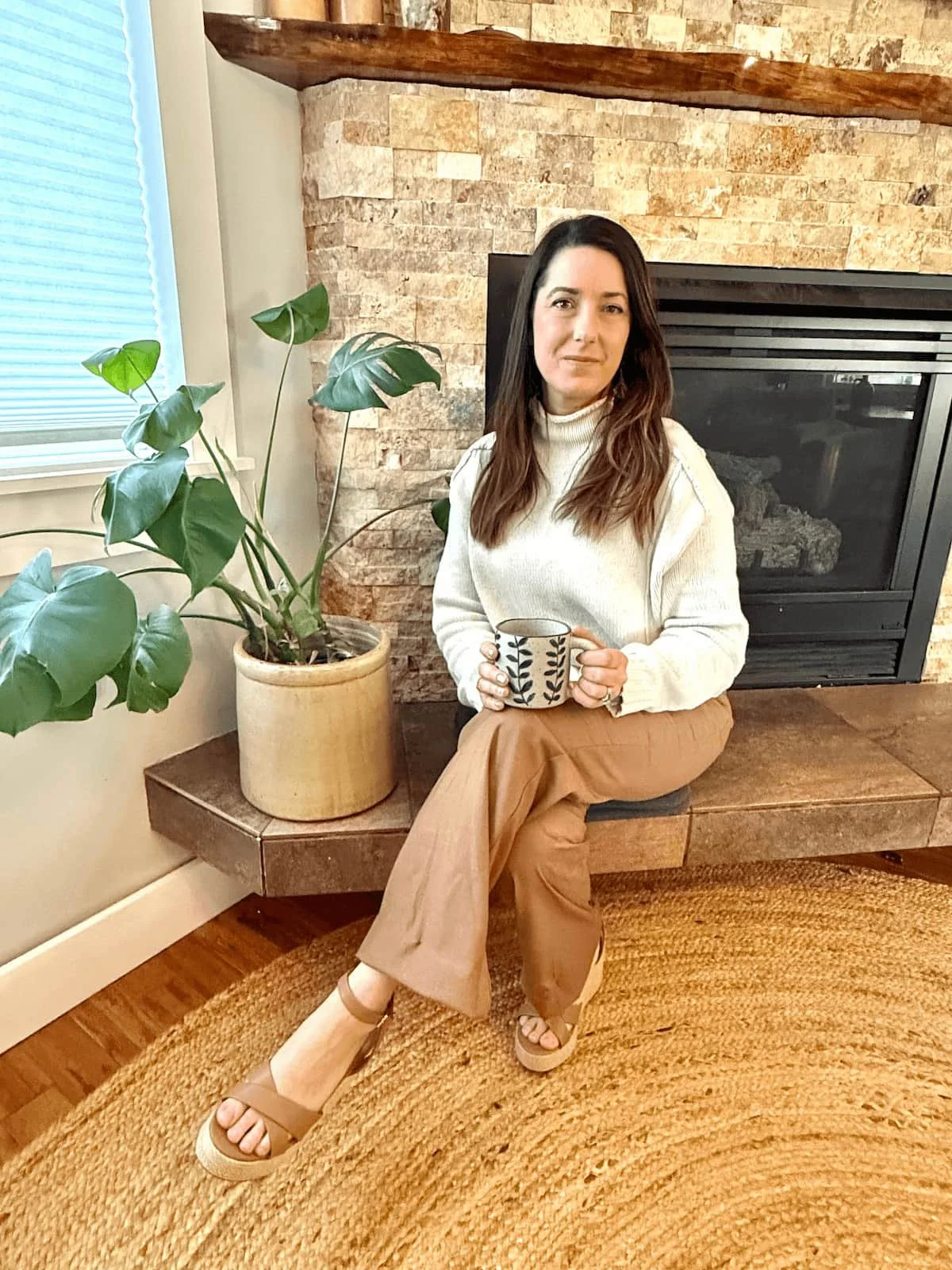 Lady with long dark hair sitting by a fireplace with a coffee inviting you to book a call