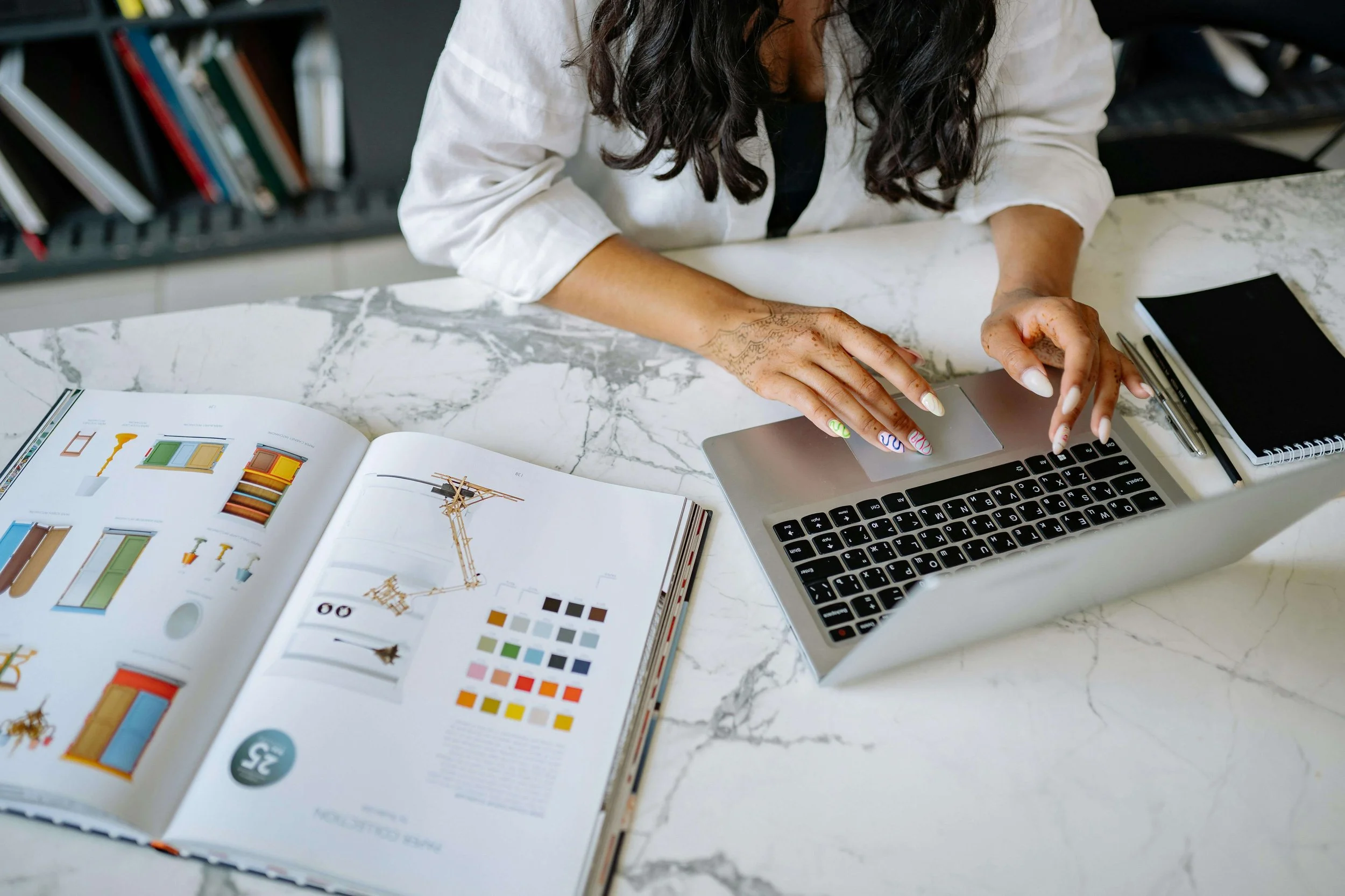 Person working at a marble table with a laptop, a book open showing design images and color palette, a black notebook, a pen, and a stylus.
