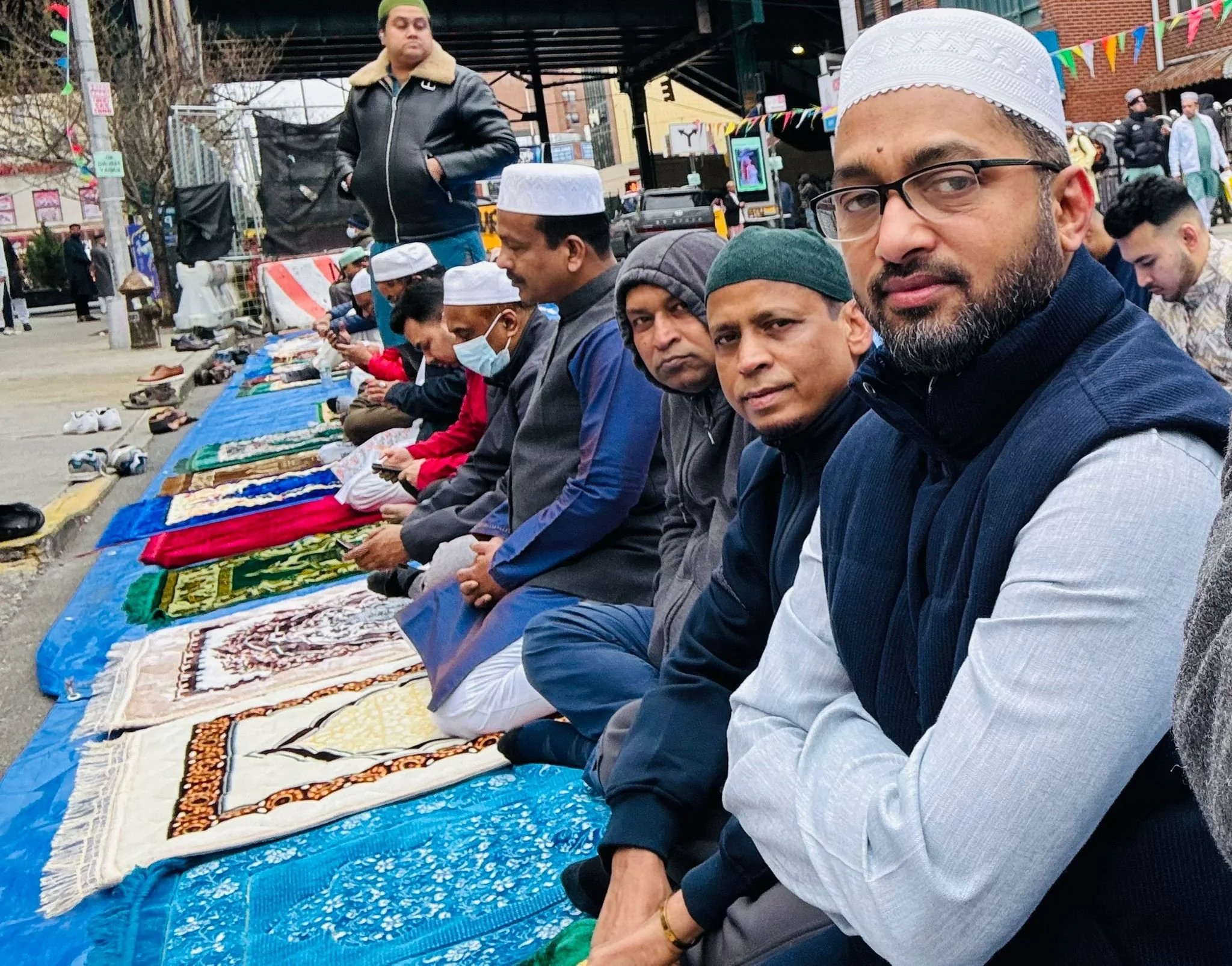 Men praying in a row on prayer rugs during an outdoor religious gathering, some wearing traditional Muslim attire and head coverings.