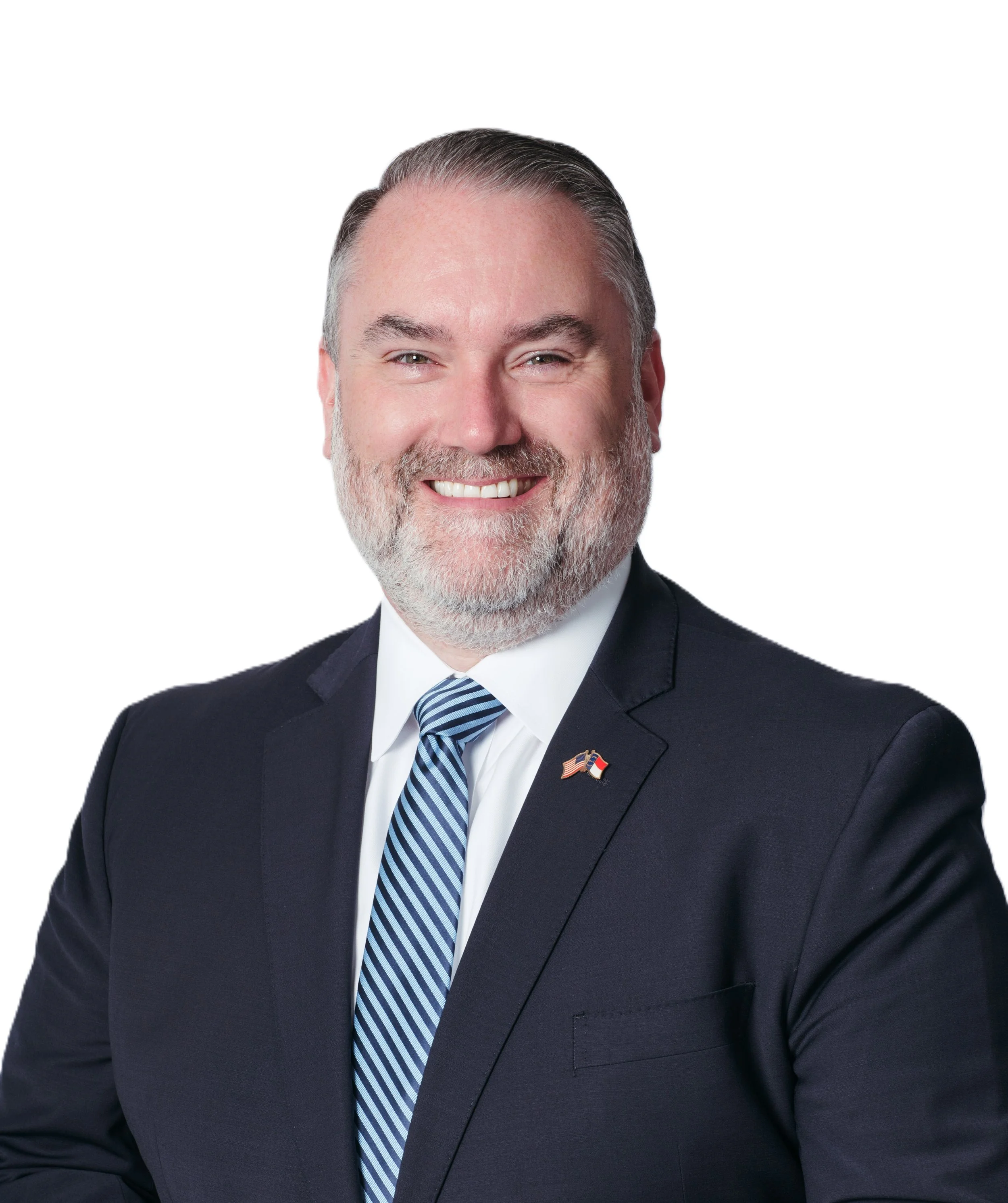 Headshot of a middle-aged man with gray hair and beard, wearing a dark suit and striped tie, against a white background, with a small American flag pin on his lapel.