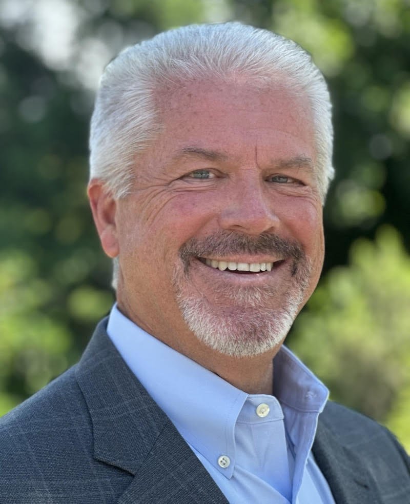 Headshot of a middle-aged man with gray hair, a beard, and a smiling expression, wearing a blue collared shirt and a dark blazer, outdoors with a blurred green leafy background.