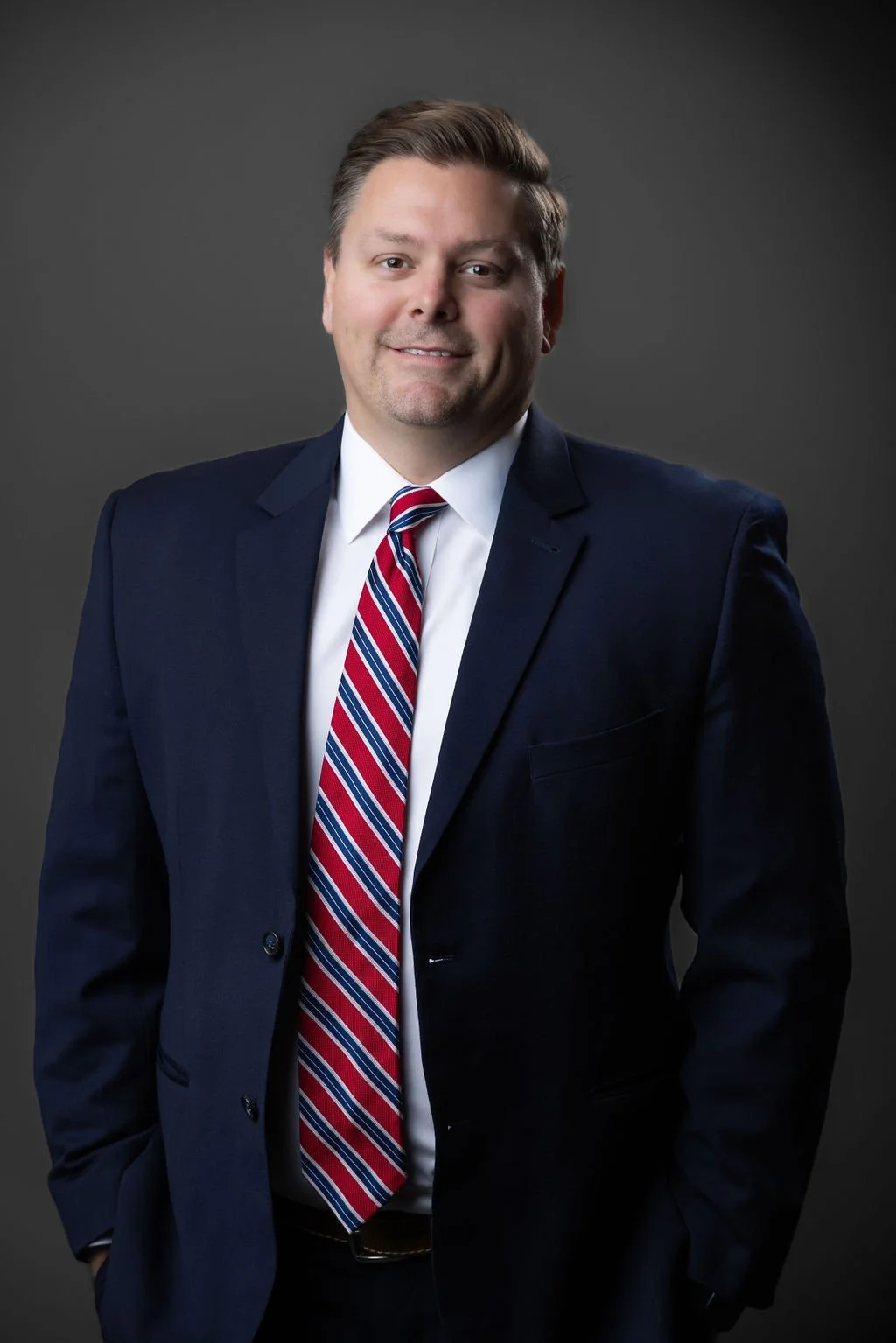 A man in a dark blue suit, white shirt, and striped red, white, and blue tie, smiling against a plain gray background.