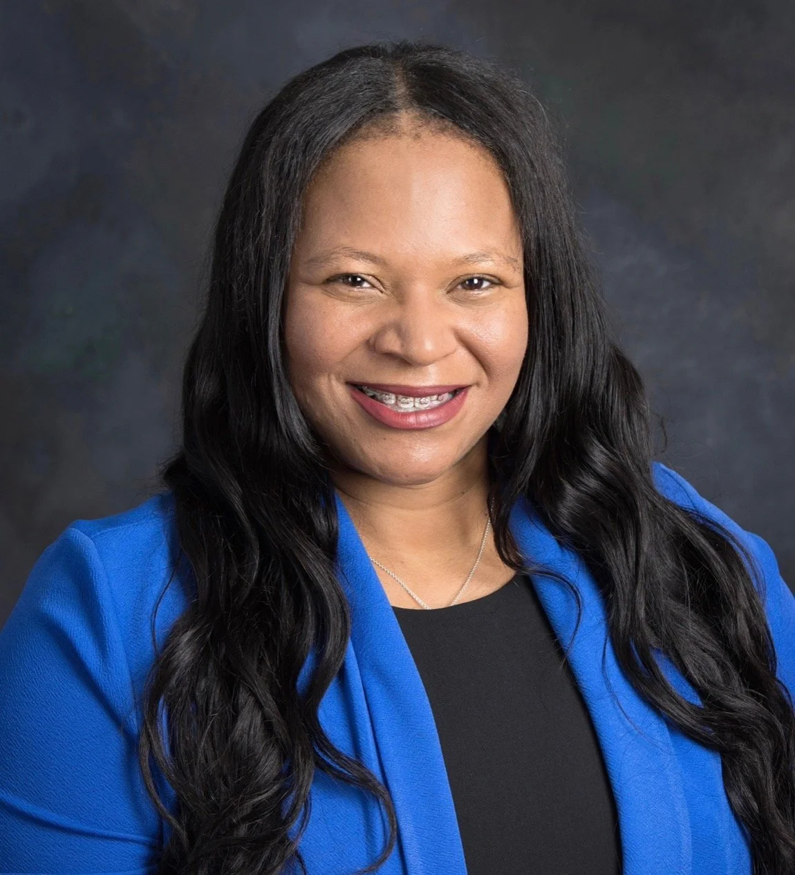 Professional headshot of a smiling woman with long black hair, wearing a blue blazer and a black top, in front of a dark background.