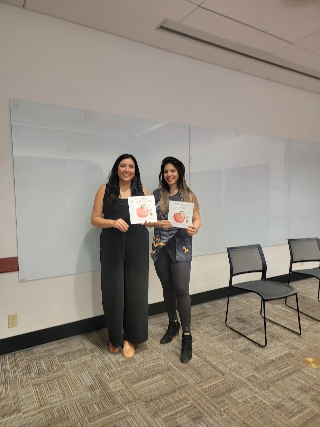 Two women standing in a room holding certificates or artwork with apple illustrations, smiling at the camera.