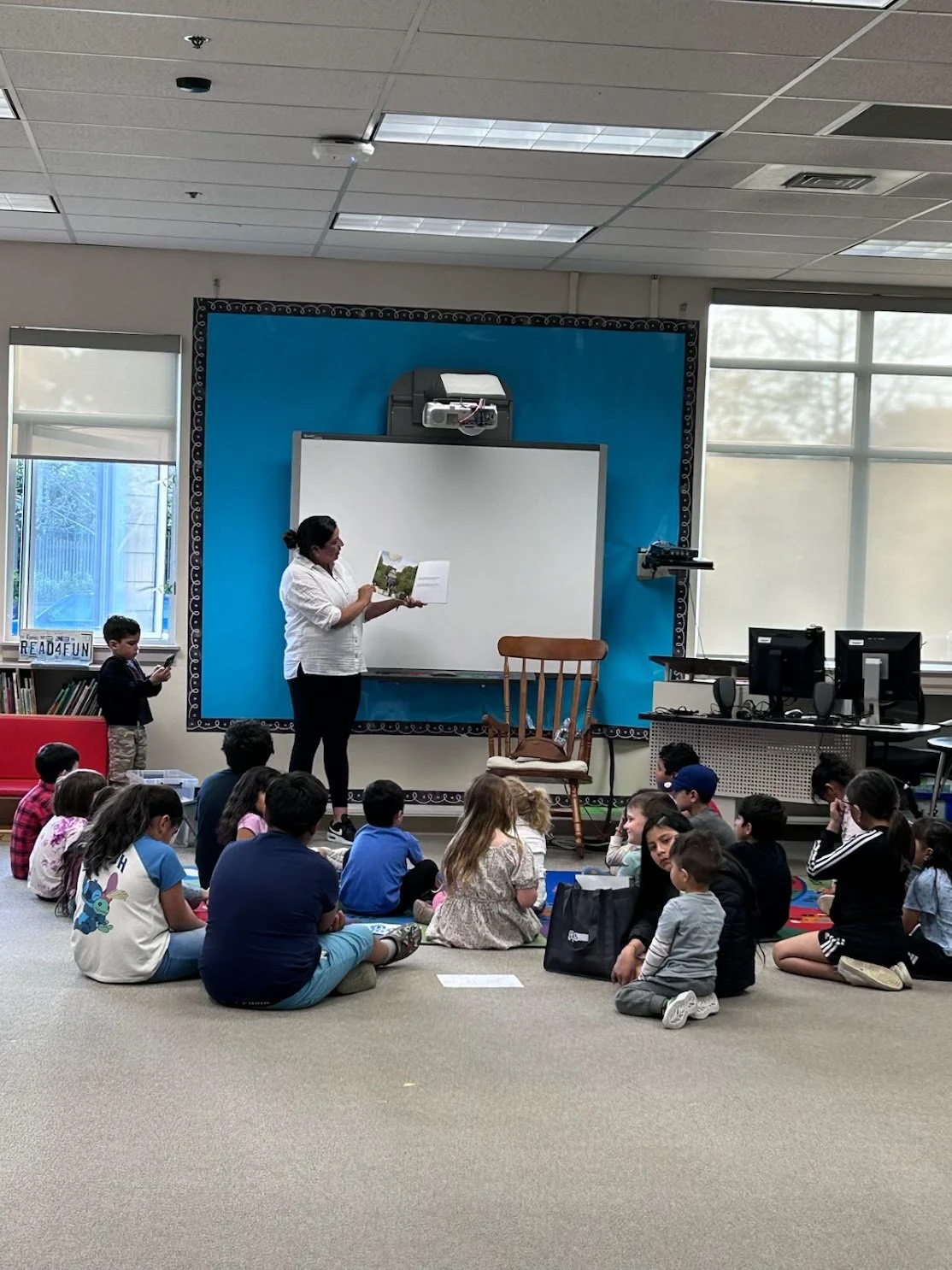 A classroom with a teacher standing in front of a whiteboard reading a book to a seated group of children. The children are sitting on the carpet, and there are windows, computers, and a rocking chair in the background.