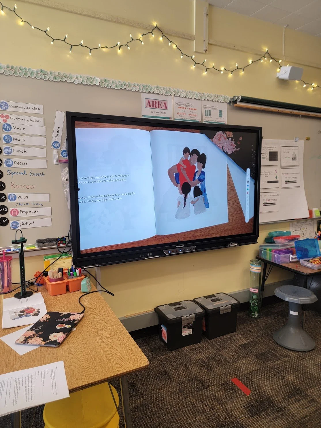 A classroom with a large digital display showing an open book with a family illustration, surrounded by classroom supplies, a whiteboard with classroom schedule notes, and decorative string lights overhead.