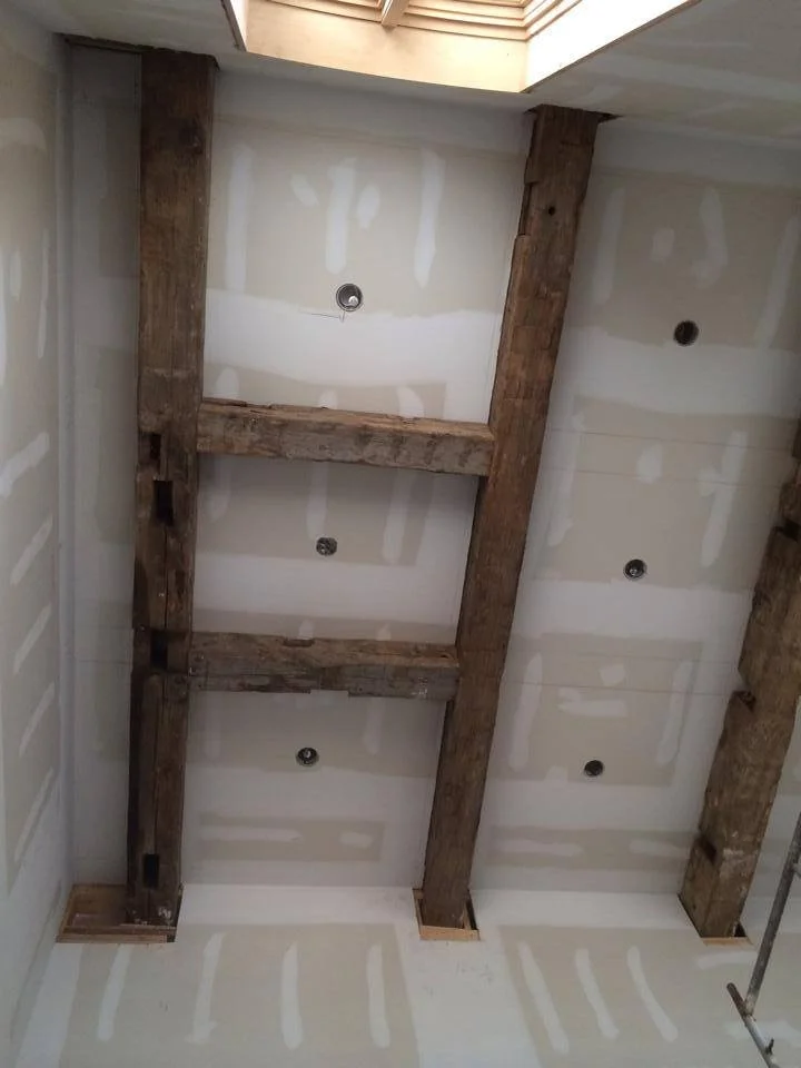 Ceiling under construction with exposed wooden beams and recessed lighting fixtures in drywall ceiling.